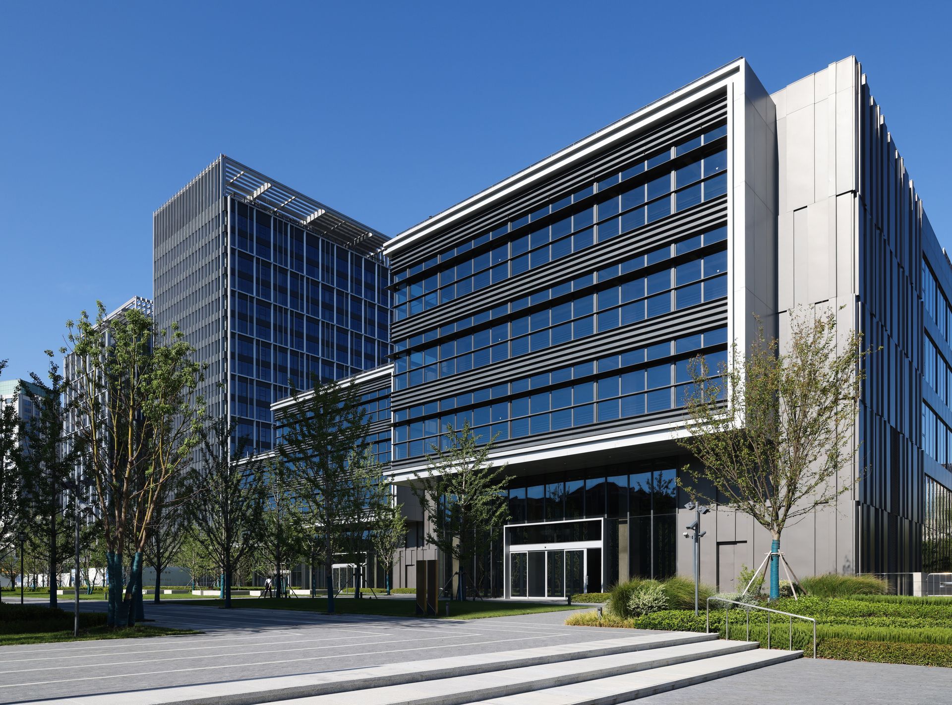 A modern office complex with glass-paneled facades, metal accents, and surrounding landscaping under a bright blue sky.