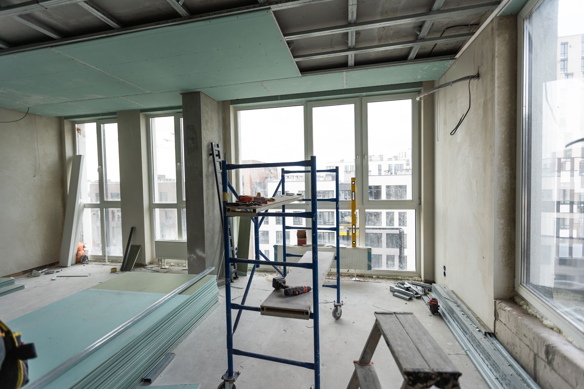 Interior of a room under construction with scaffolding, plasterboard sheets on the floor, and unfinished walls.