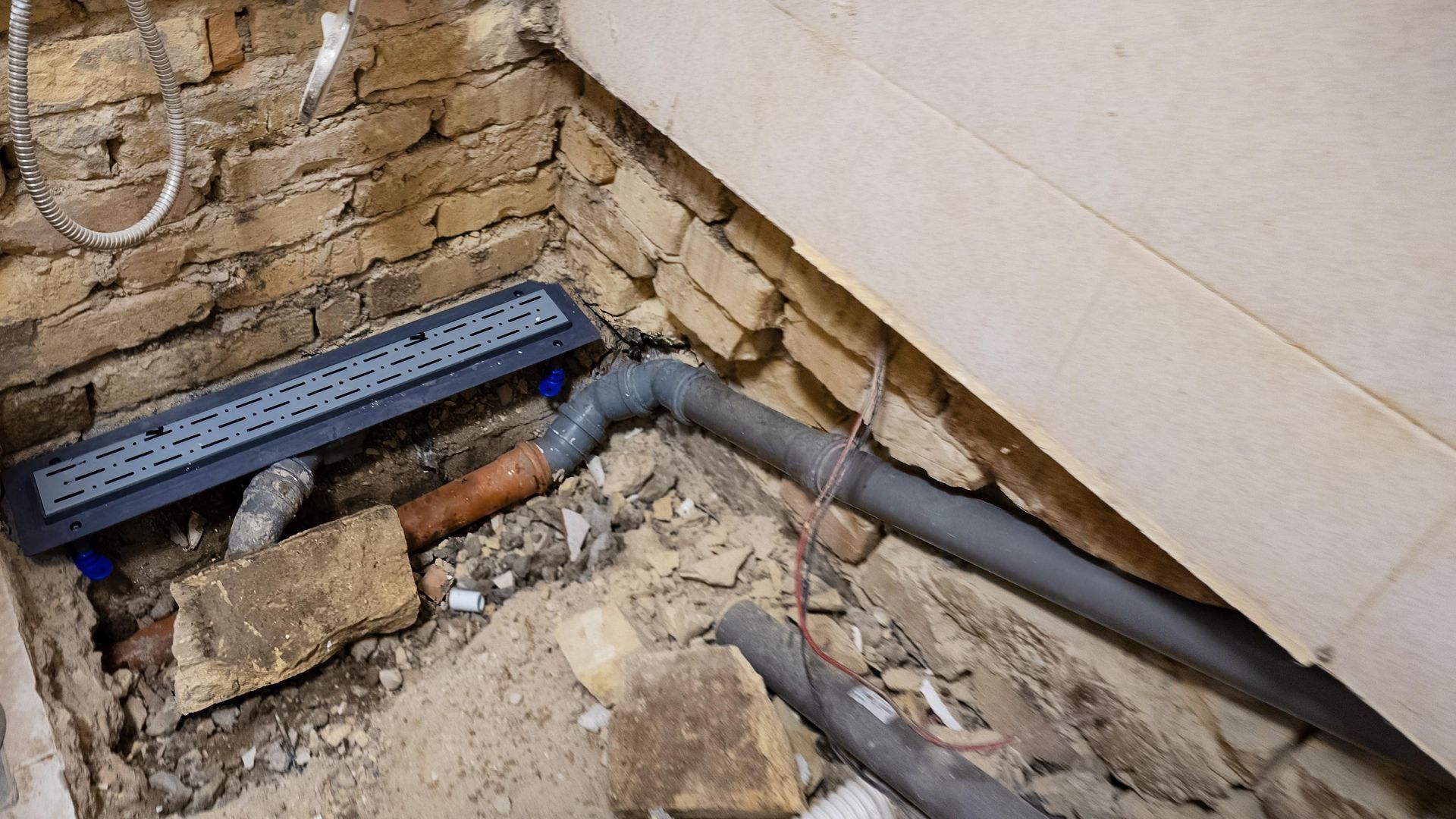 A new linear shower drain installed on a dirt floor with gray PVC pipes connecting to existing plumbing near a brick wall.