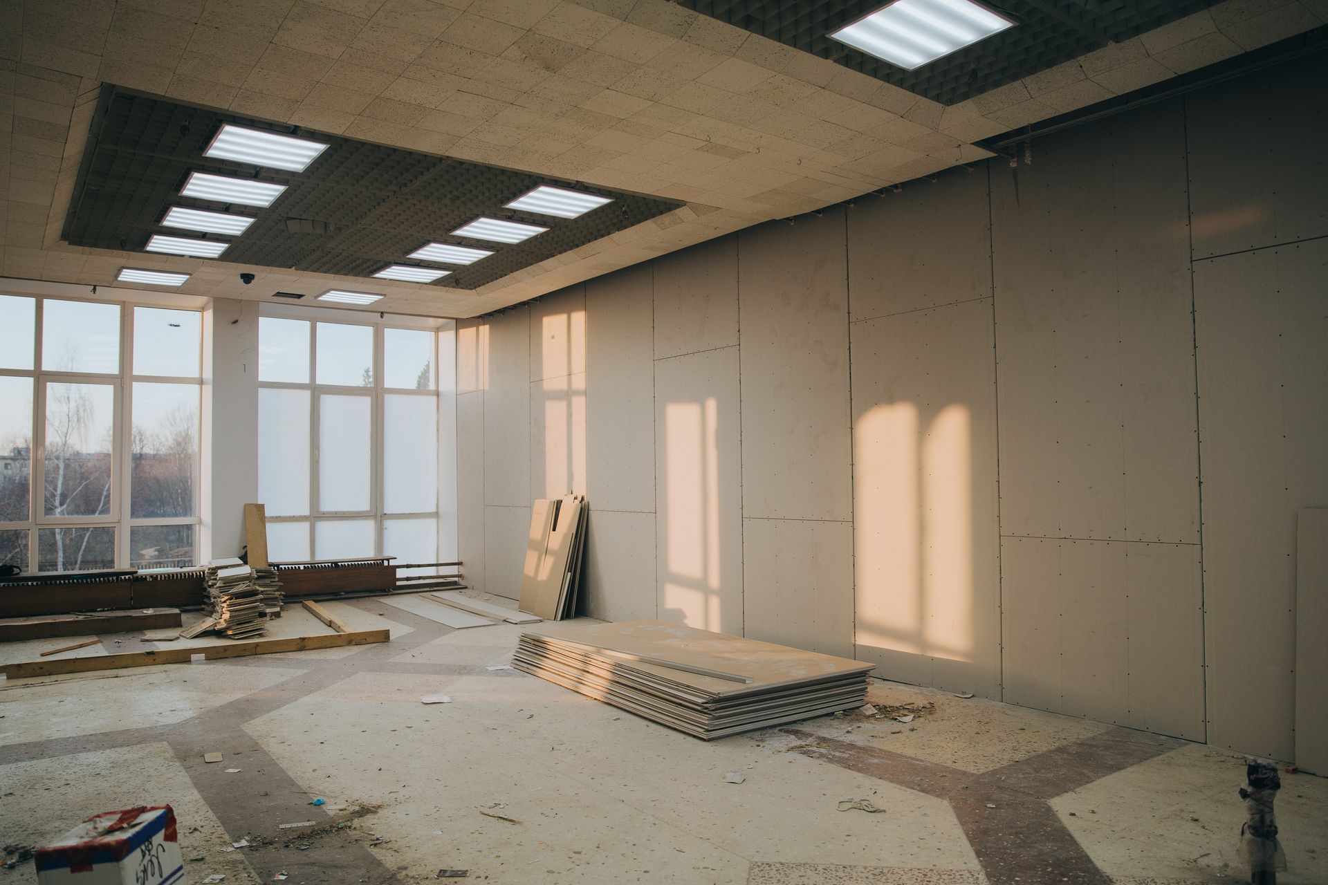 An interior view of a room under renovation with drywall walls, large windows, ceiling lights, and construction debris.