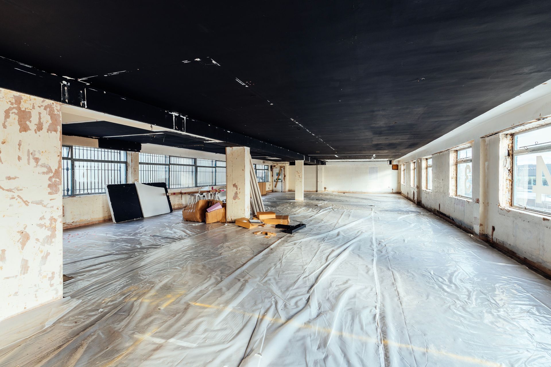A large, empty industrial space undergoing renovation with a black ceiling, white walls, and plastic-covered floors.