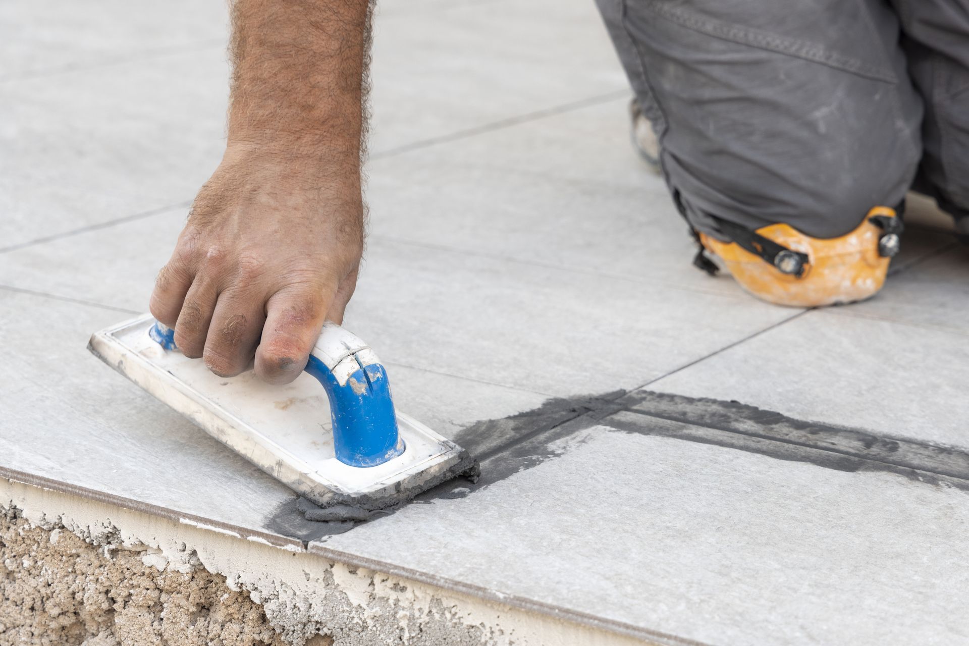 A person uses a blue and white grout float to press dark grout into the joints between light grey floor tiles.