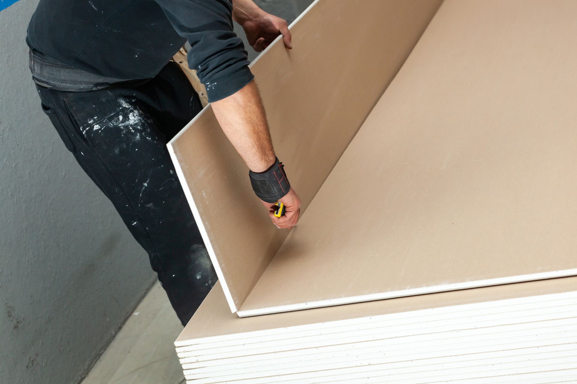 A worker in dark clothing uses a utility knife to cut a large sheet of drywall resting on a stack of boards.