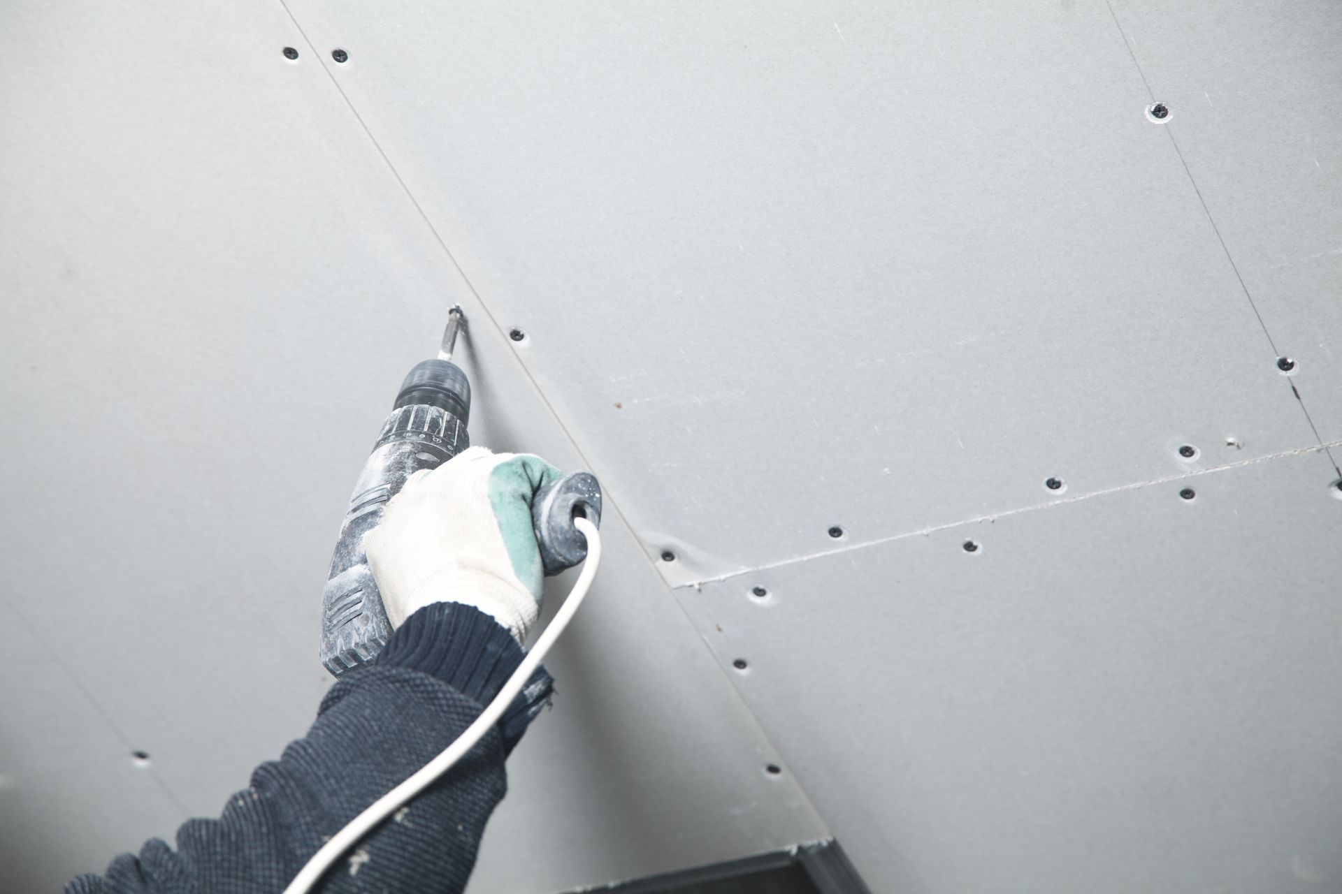 A person using a drill to fasten white drywall panels to a ceiling.