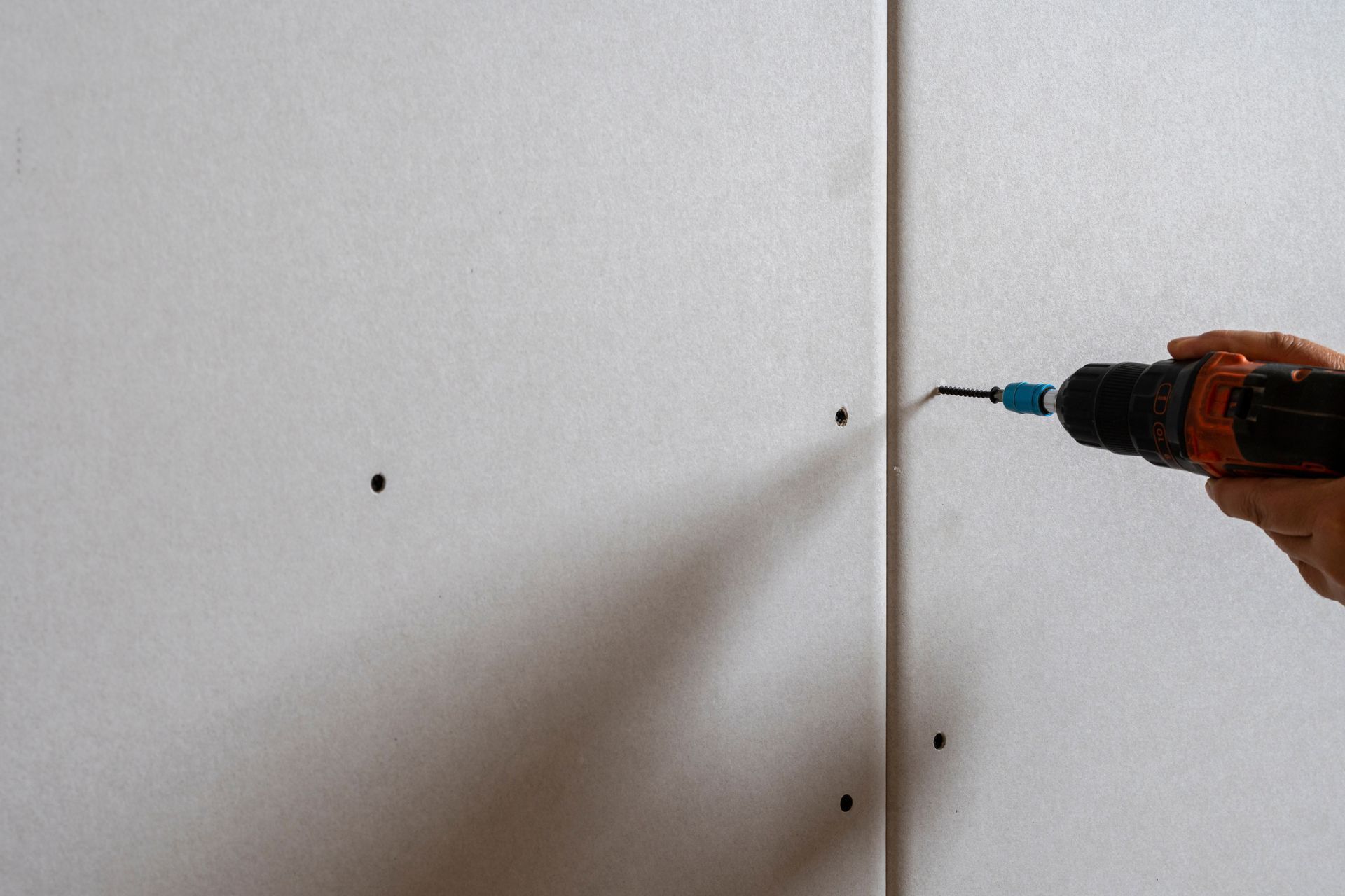 A hand uses a power drill to drive a screw into a drywall seam where two panels meet.