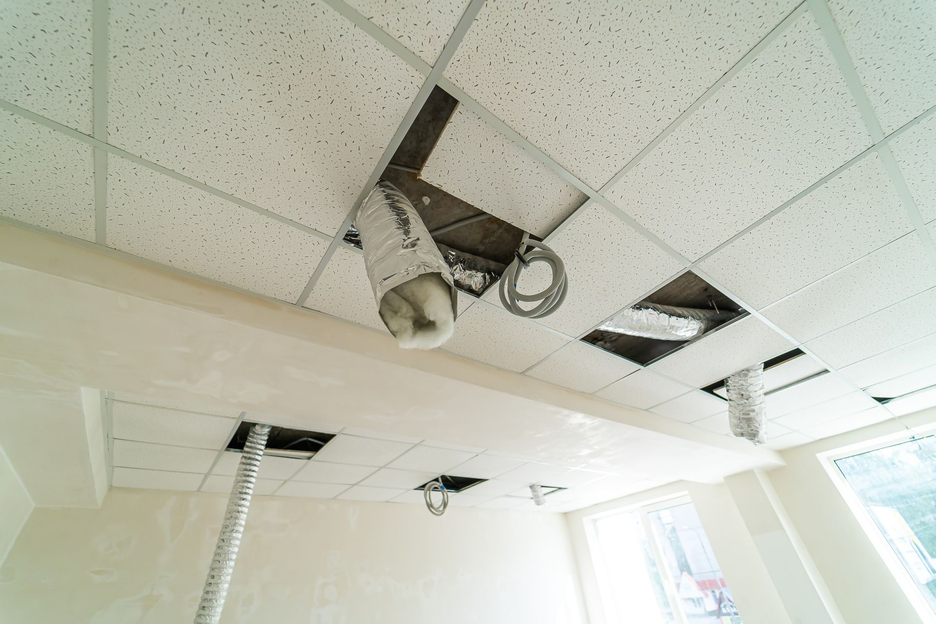 A room under renovation with an incomplete drop ceiling, exposed metal grid, wires, and flexible ventilation ducts.