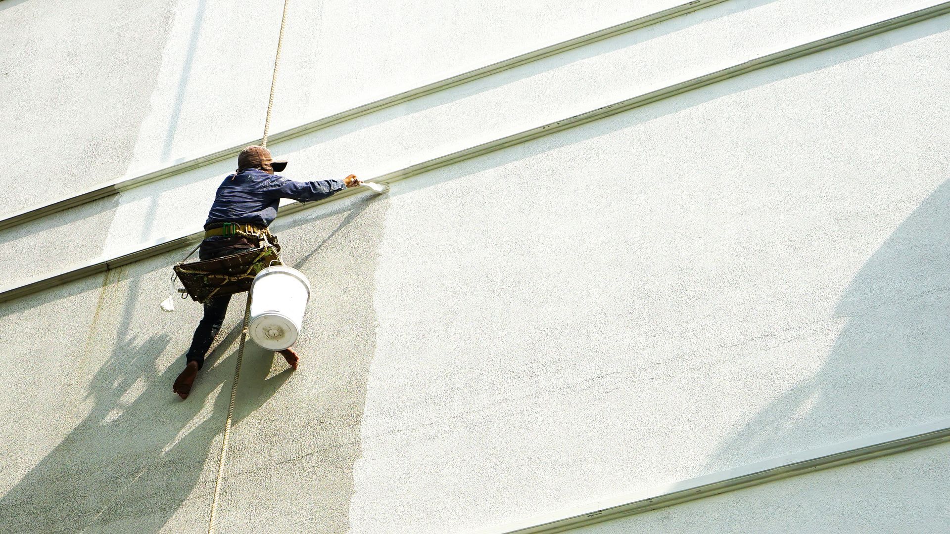 A worker suspended by ropes paints a white textured wall with a brush, carrying a white bucket.
