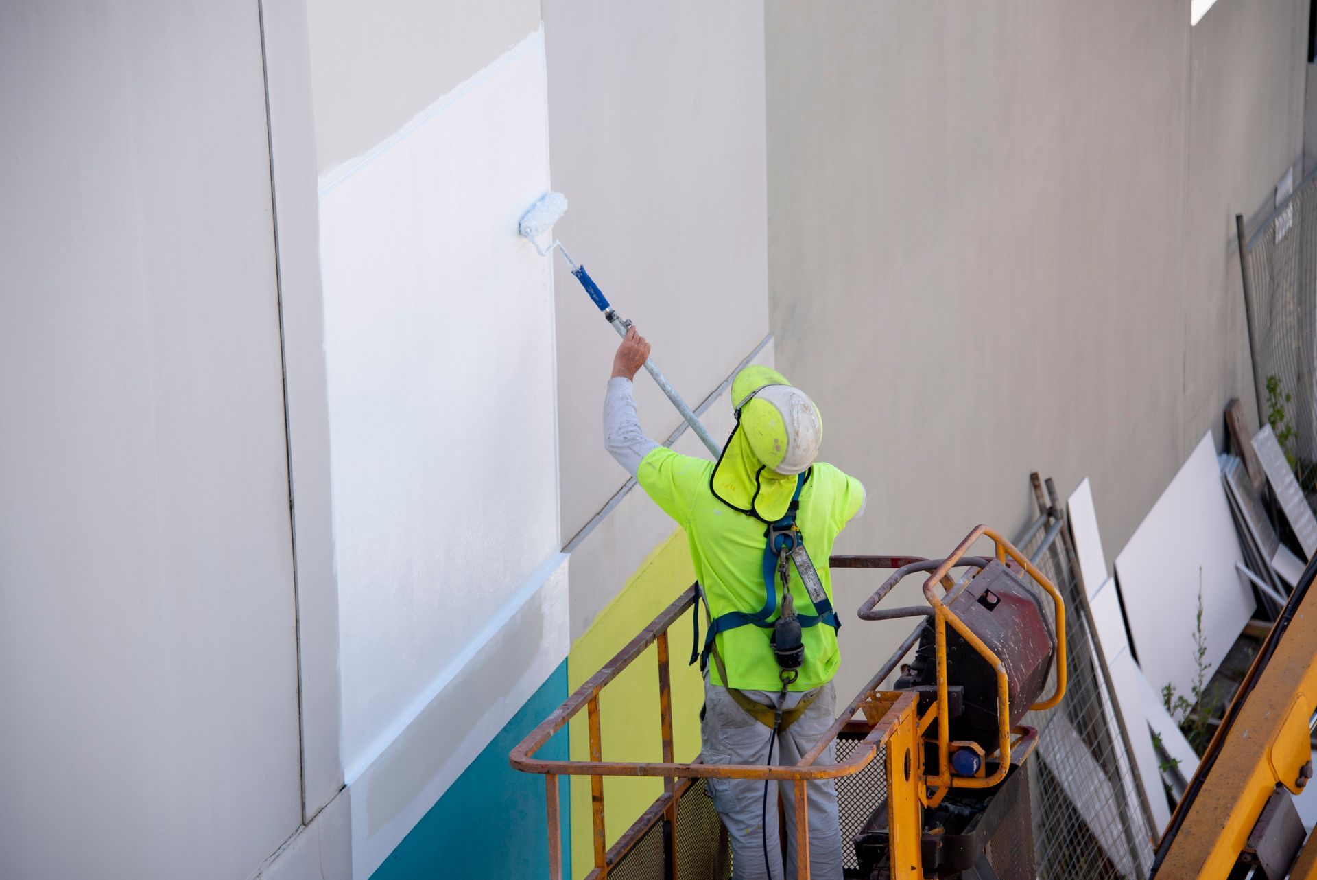 A worker in a high-visibility vest and hard hat uses a paint roller on a long extension pole to paint a wall white.