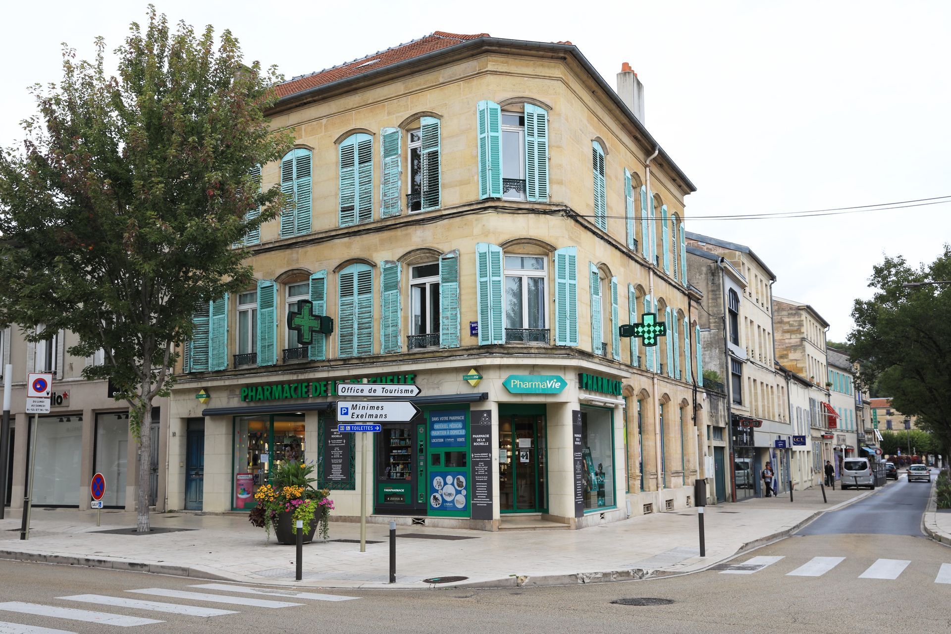A three-story corner building with a pharmacy on the ground floor, featuring light stone walls and blue shutters.