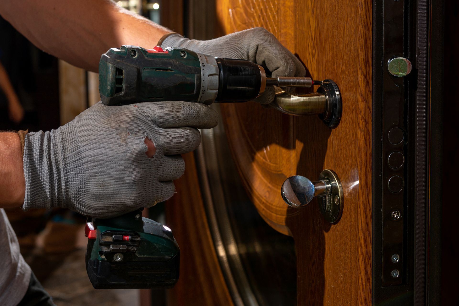 A person wearing grey work gloves uses a cordless drill to tighten a screw on a wooden door handle.