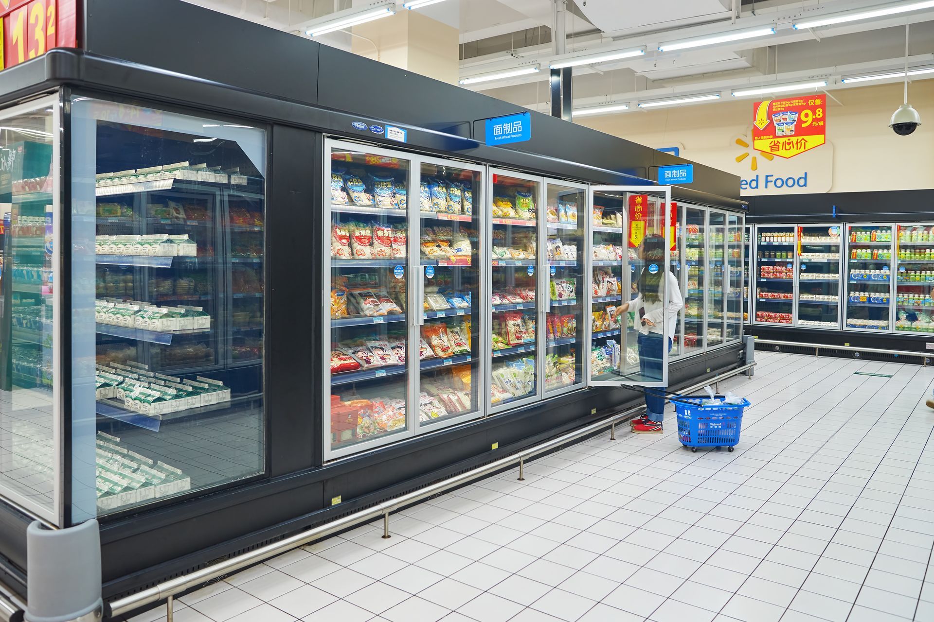 A shopper stands by glass-door freezer cases in a well-lit grocery store, holding a blue basket.
