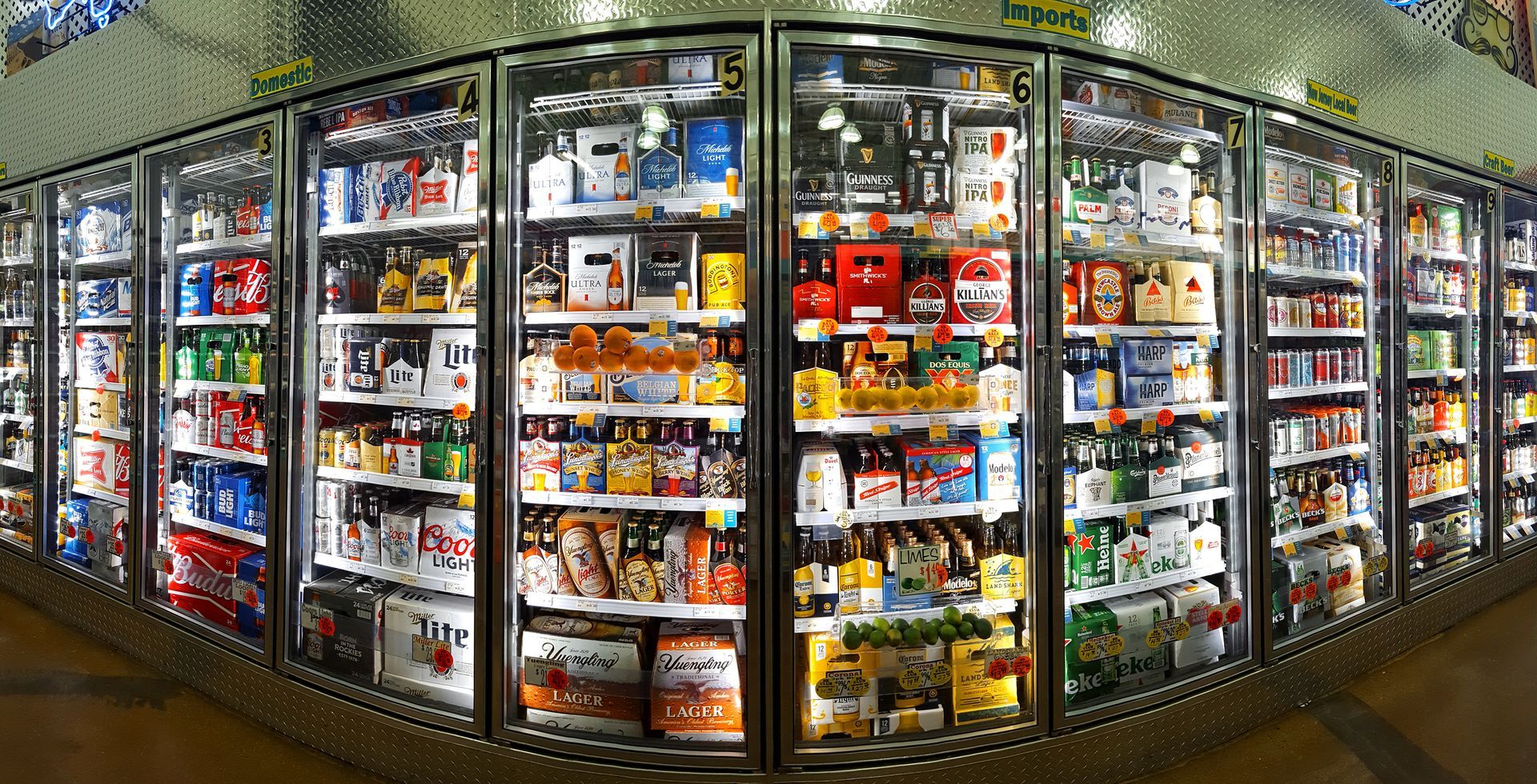 A wide shot of a refrigerated beer aisle in a store, featuring multiple glass-door coolers filled with various brands.