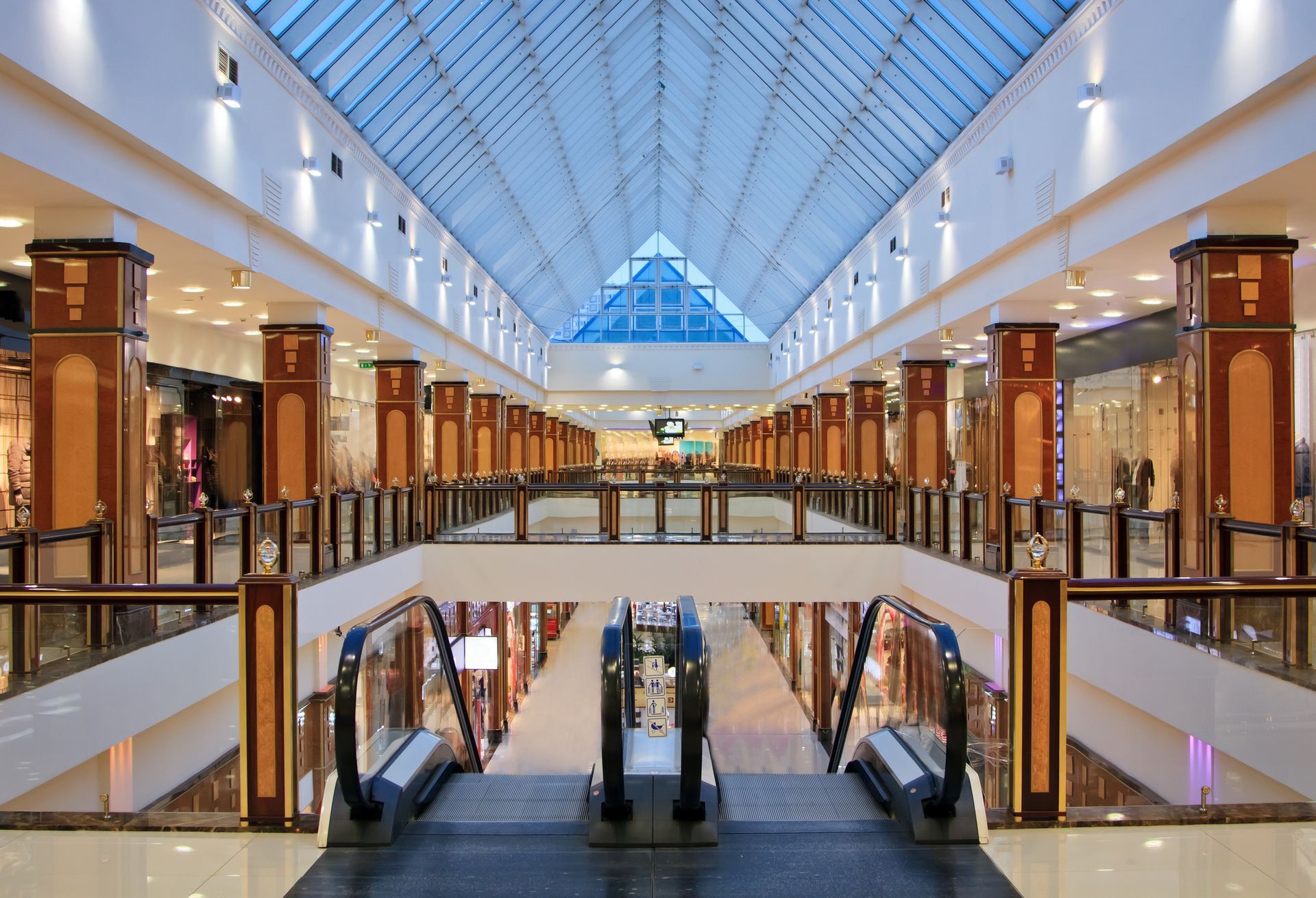 A symmetrical, modern shopping mall interior featuring a glass atrium ceiling, dark wood columns, and central escalators.