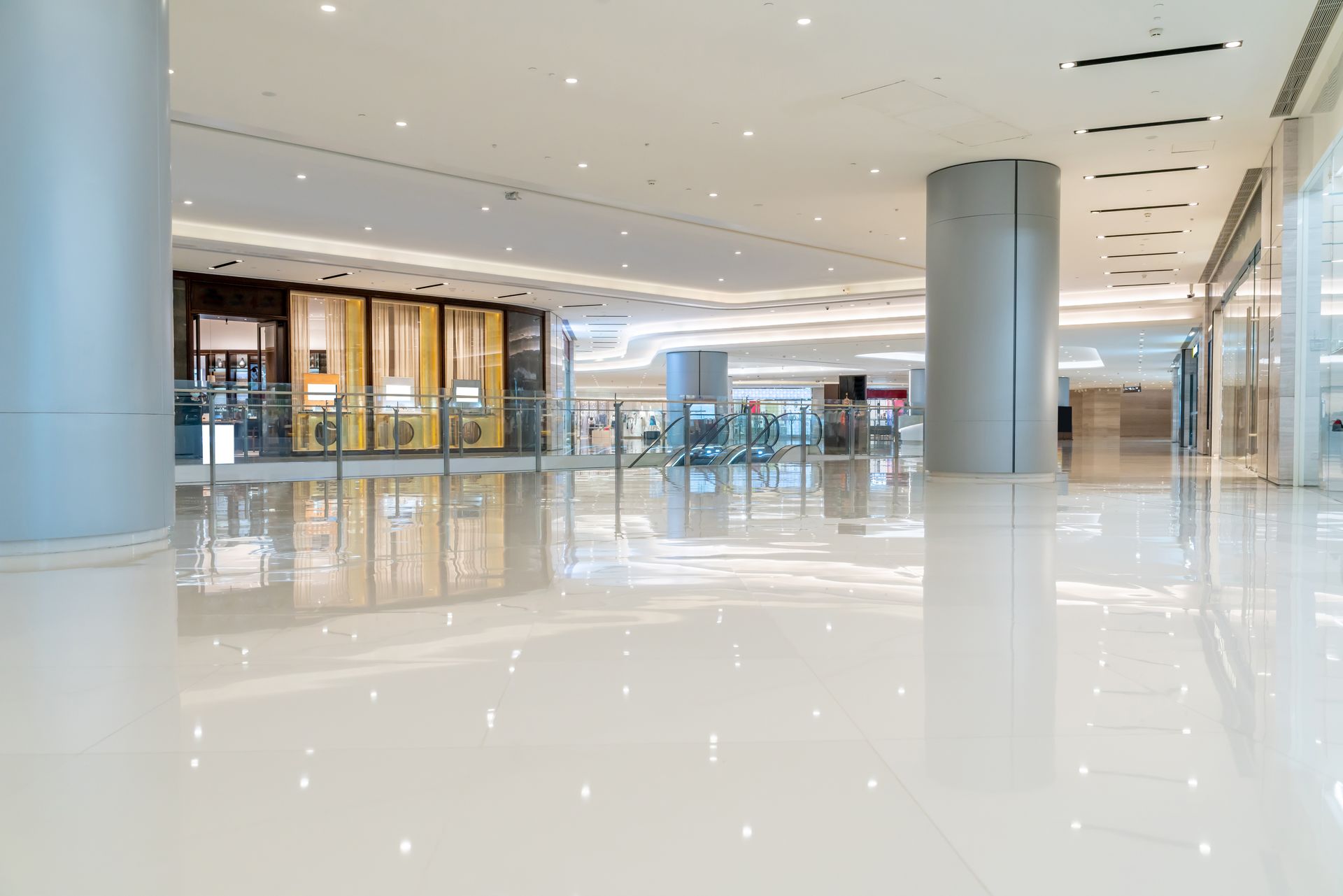 A wide, empty, modern shopping mall hallway with polished white floors, recessed lighting, and large cylindrical pillars.