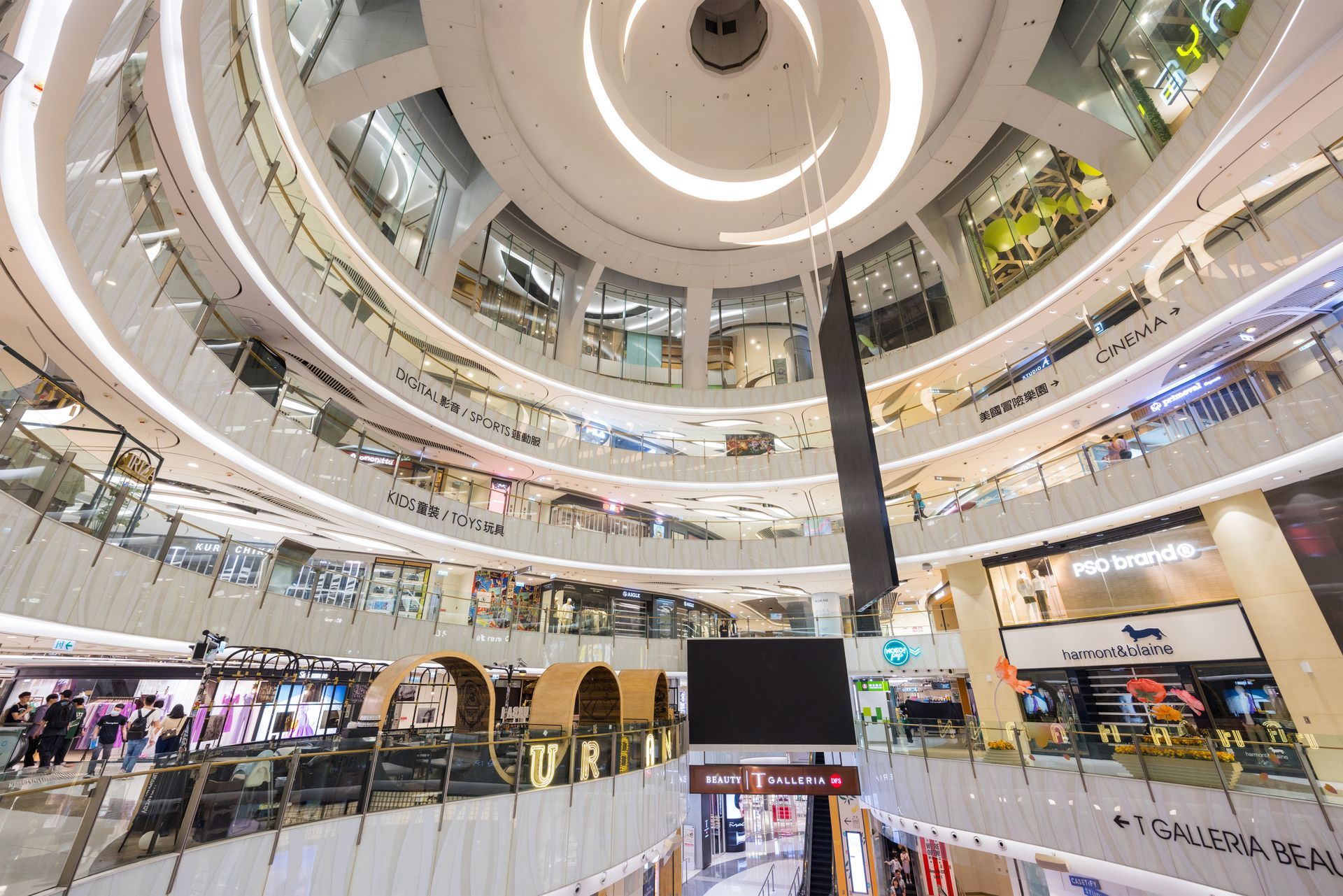 A multi-level shopping mall atrium with a circular skylight, curved balconies, glass railings, and various storefronts.