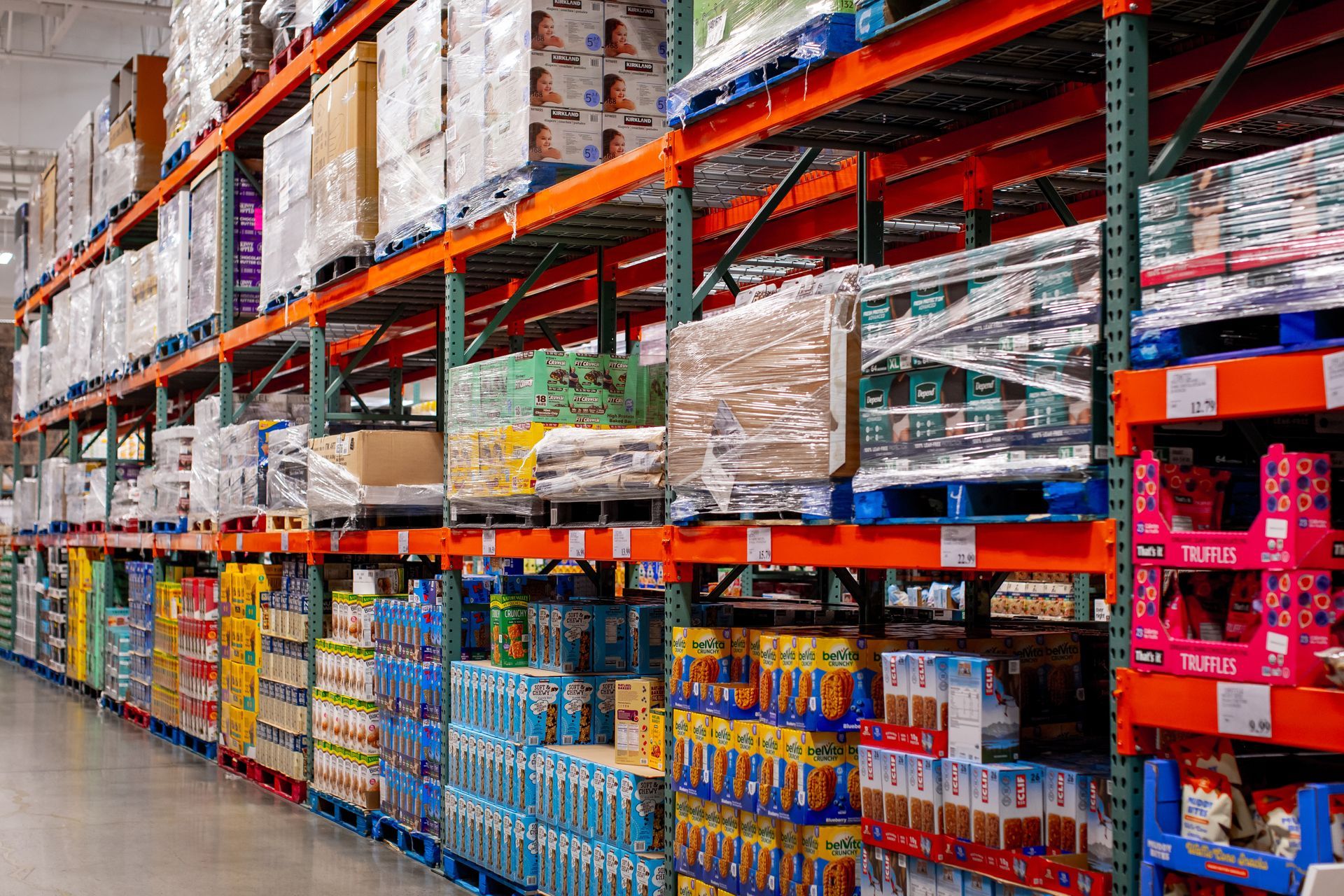 A view down a warehouse aisle with tall orange metal shelves stocked with bulk goods and products on pallets.