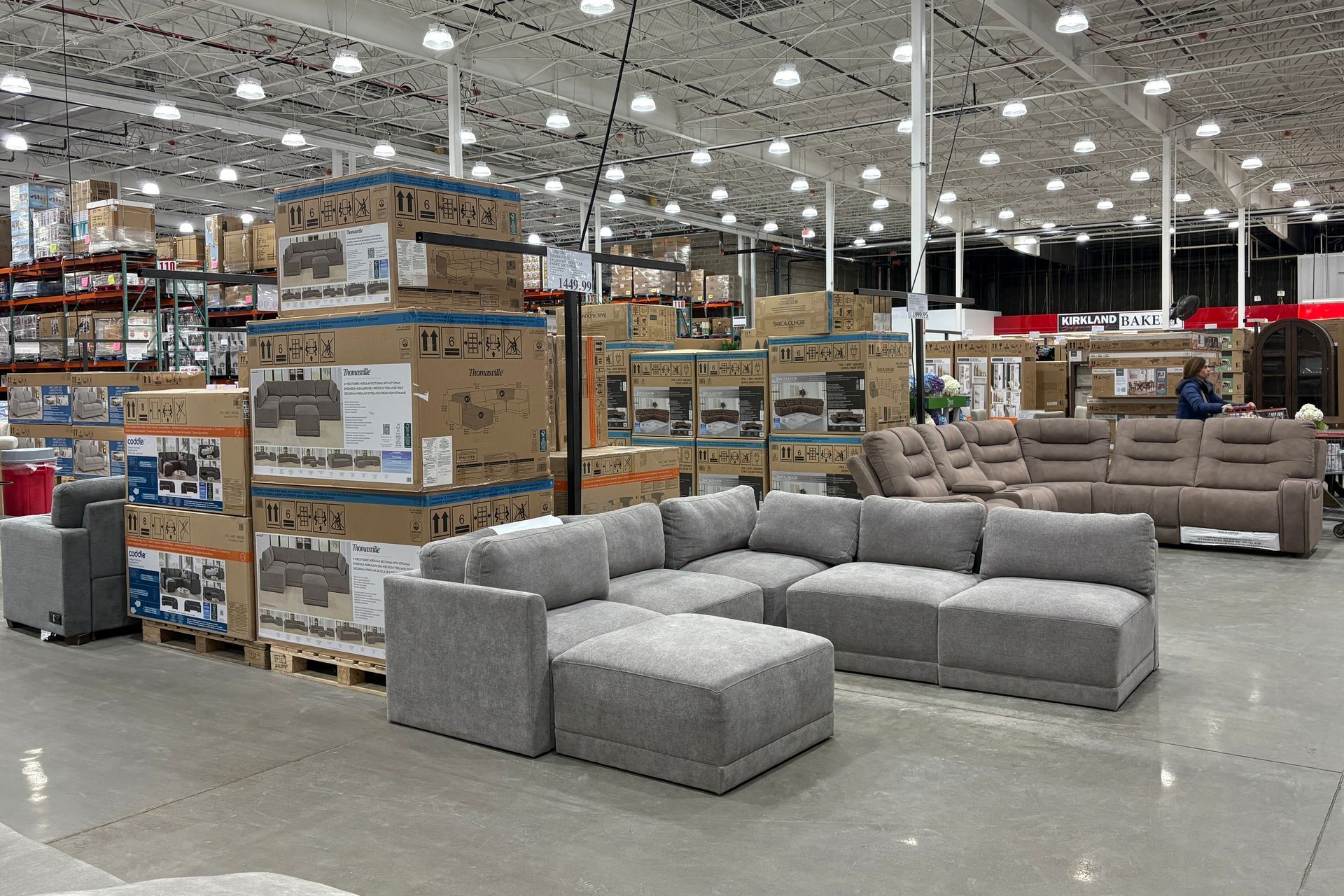 Sectional couches displayed for sale in a warehouse store with stacks of boxed furniture in the background.