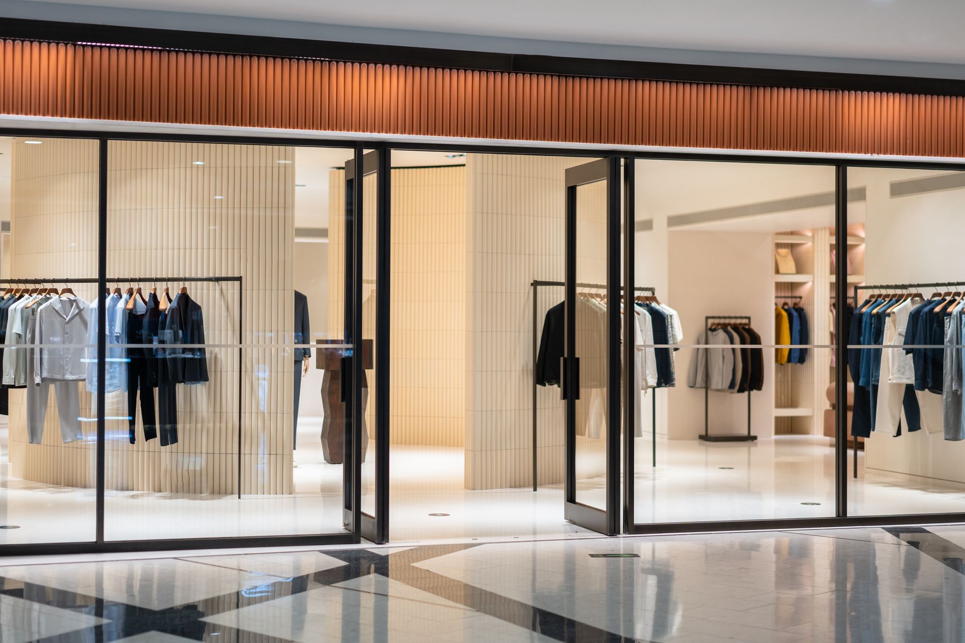 A storefront entrance to a modern clothing boutique with neutral decor, glass doors, and racks of organized apparel.