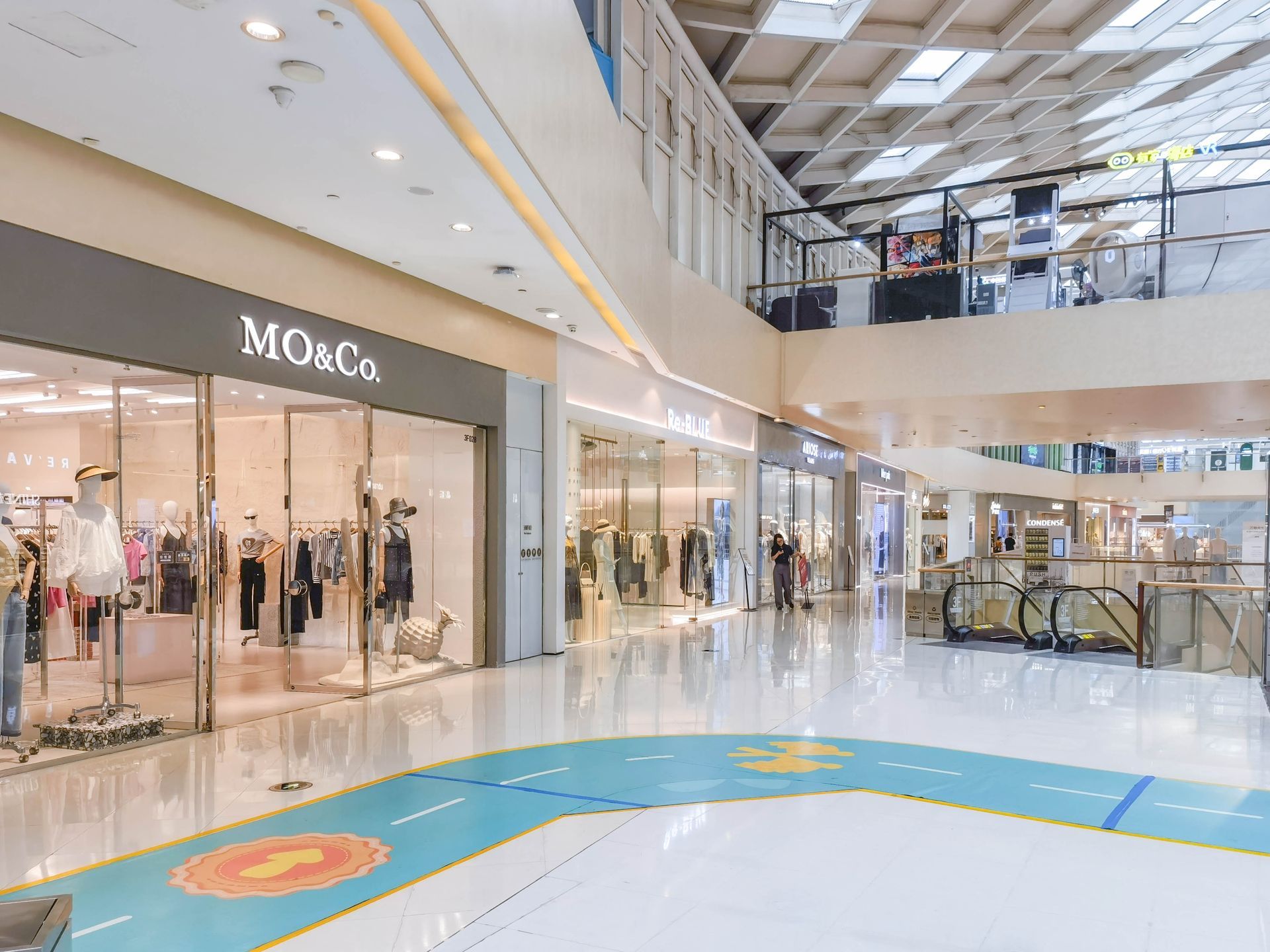 A wide shot of a modern, brightly lit shopping mall corridor with clothing storefronts and a graphic floor decal.