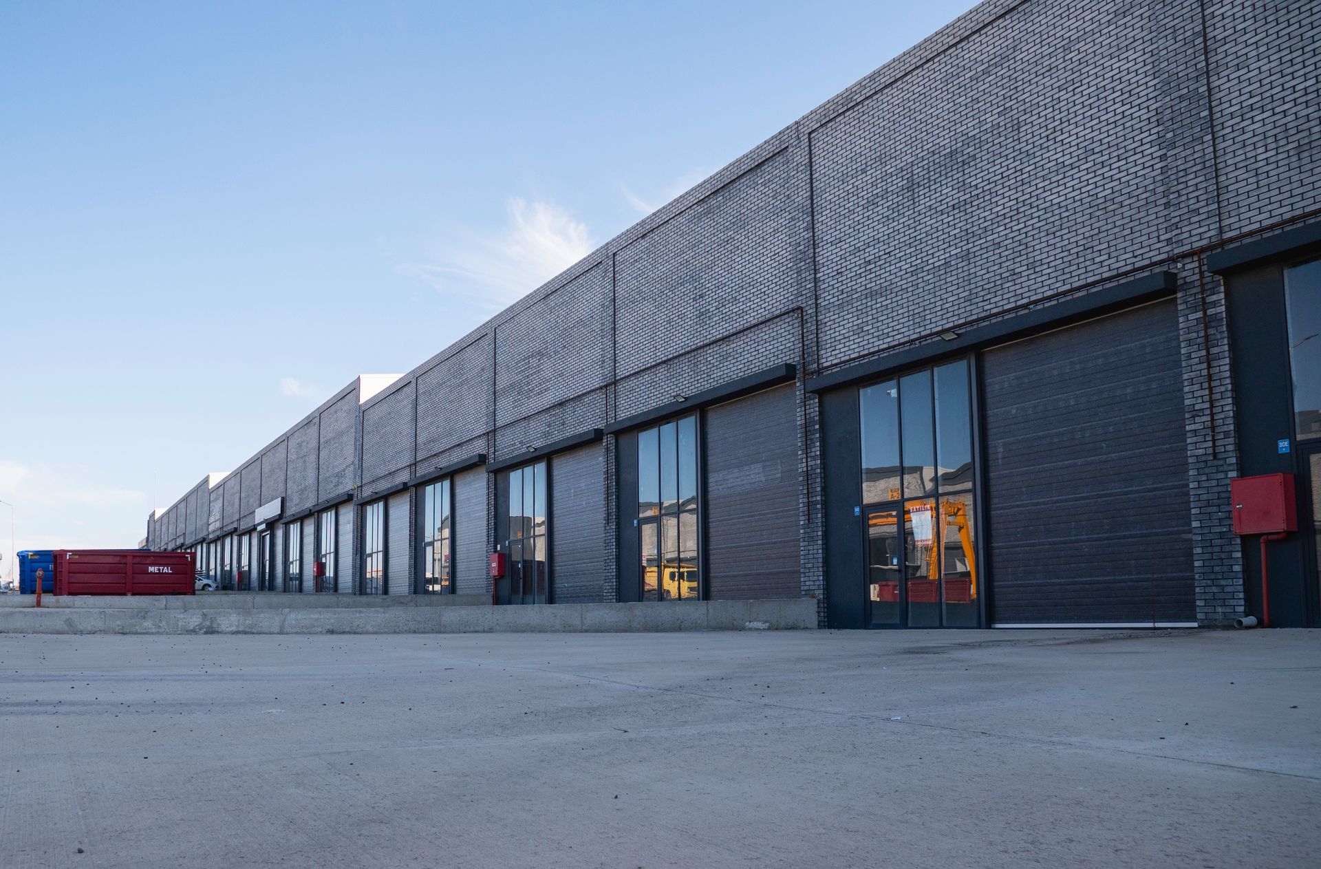 A low-angle view of a long, single-story industrial building featuring dark, textured brick walls and rows of glass doors.