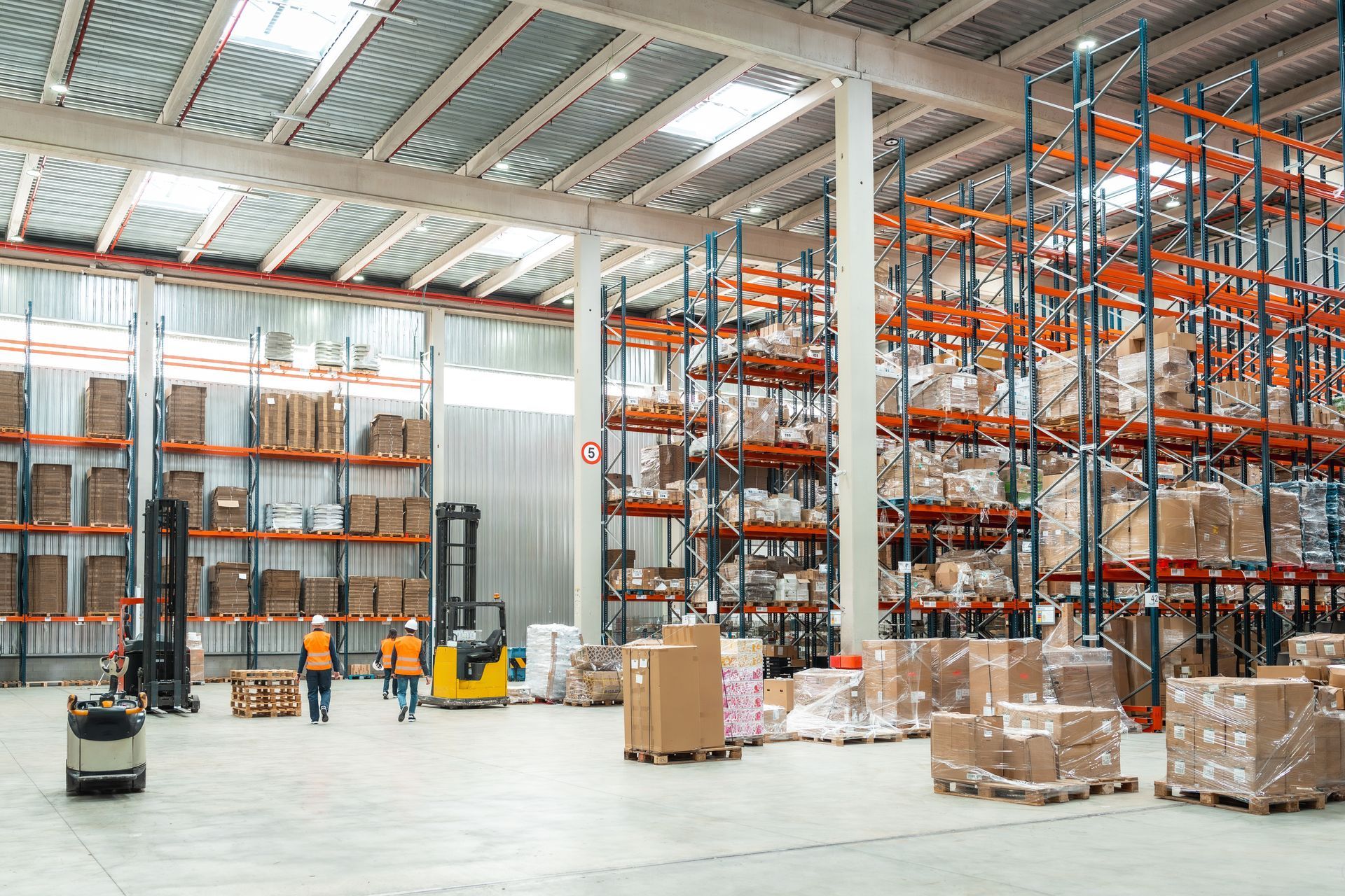 Two warehouse workers in orange vests walk through a large facility filled with tall, loaded storage racks and forklifts.