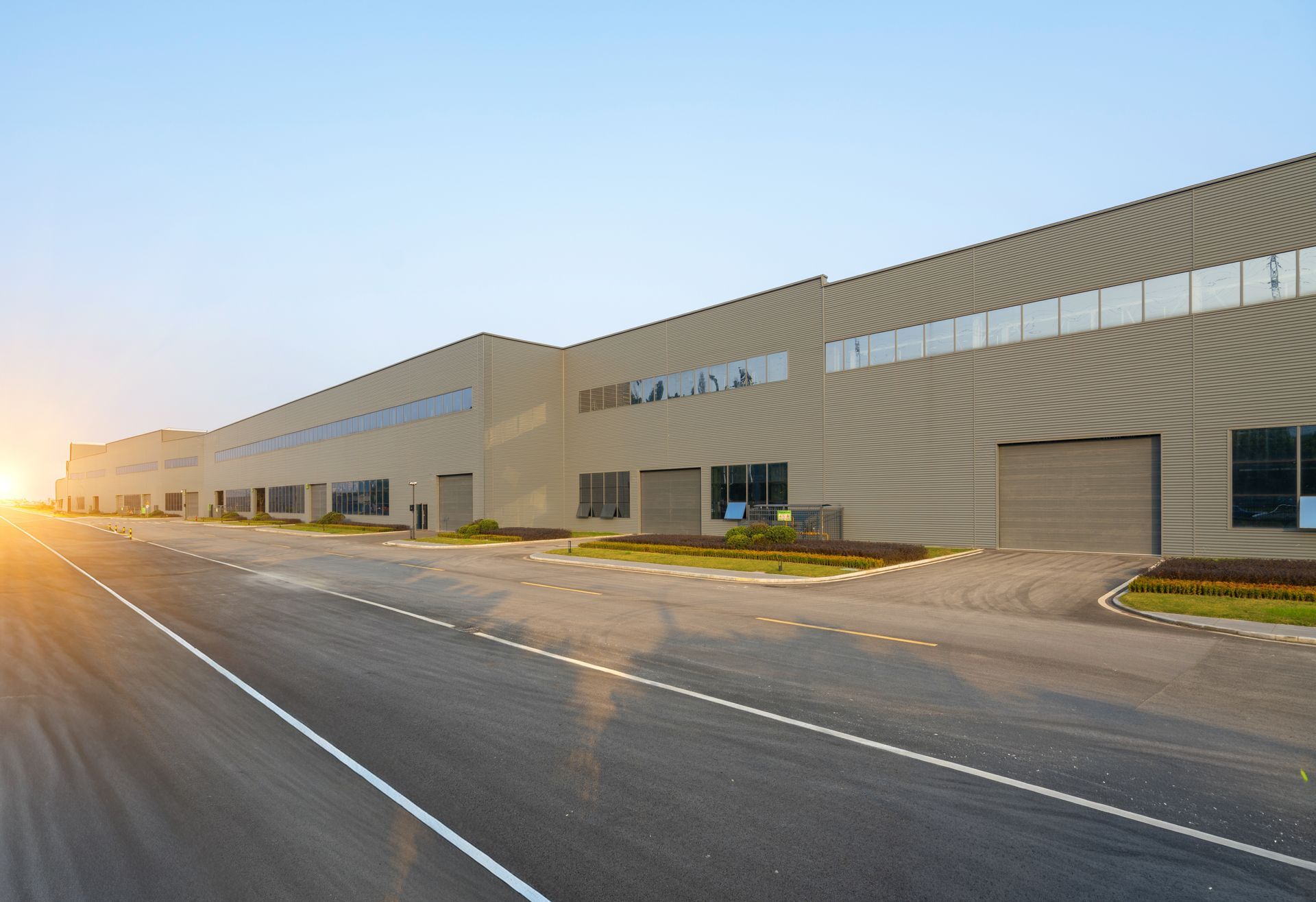 A long, industrial warehouse building with metal siding sits beside a paved road under a clear sky at sunset.