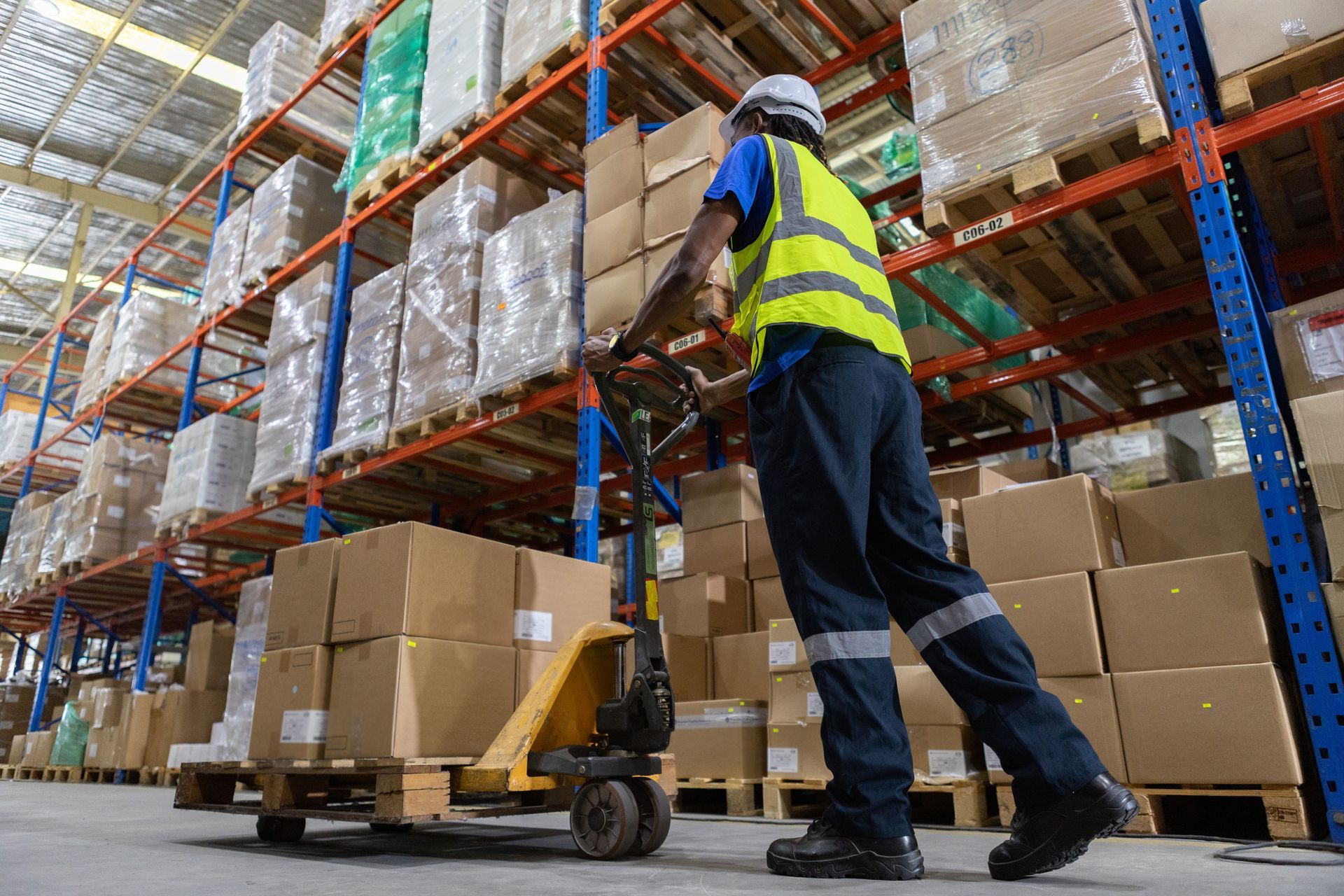 A warehouse worker in a high-visibility vest pulls a pallet jack loaded with cardboard boxes through an aisle.