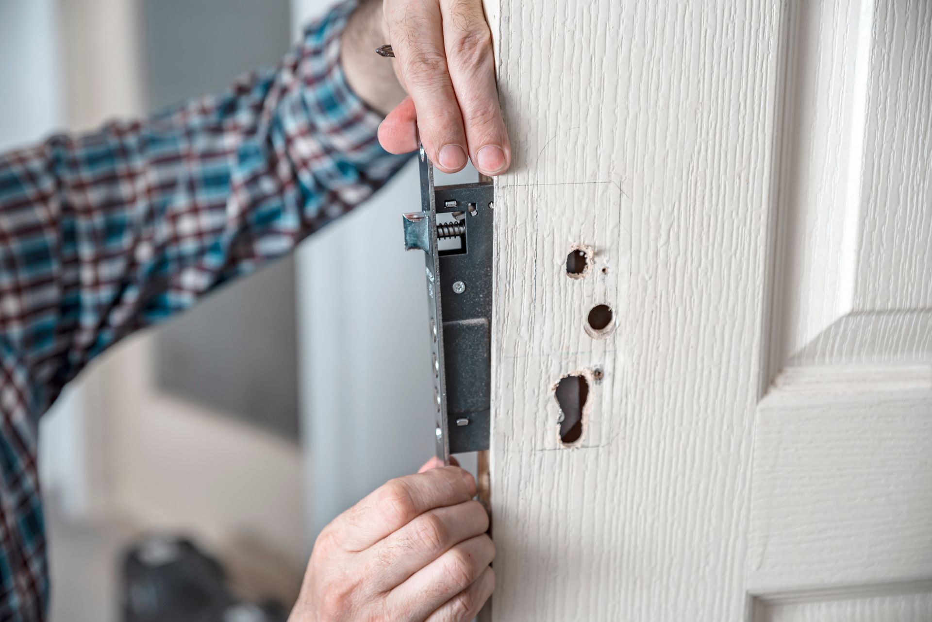 A person in a plaid shirt installs a metal mortise lock mechanism into the side of a white wooden door.