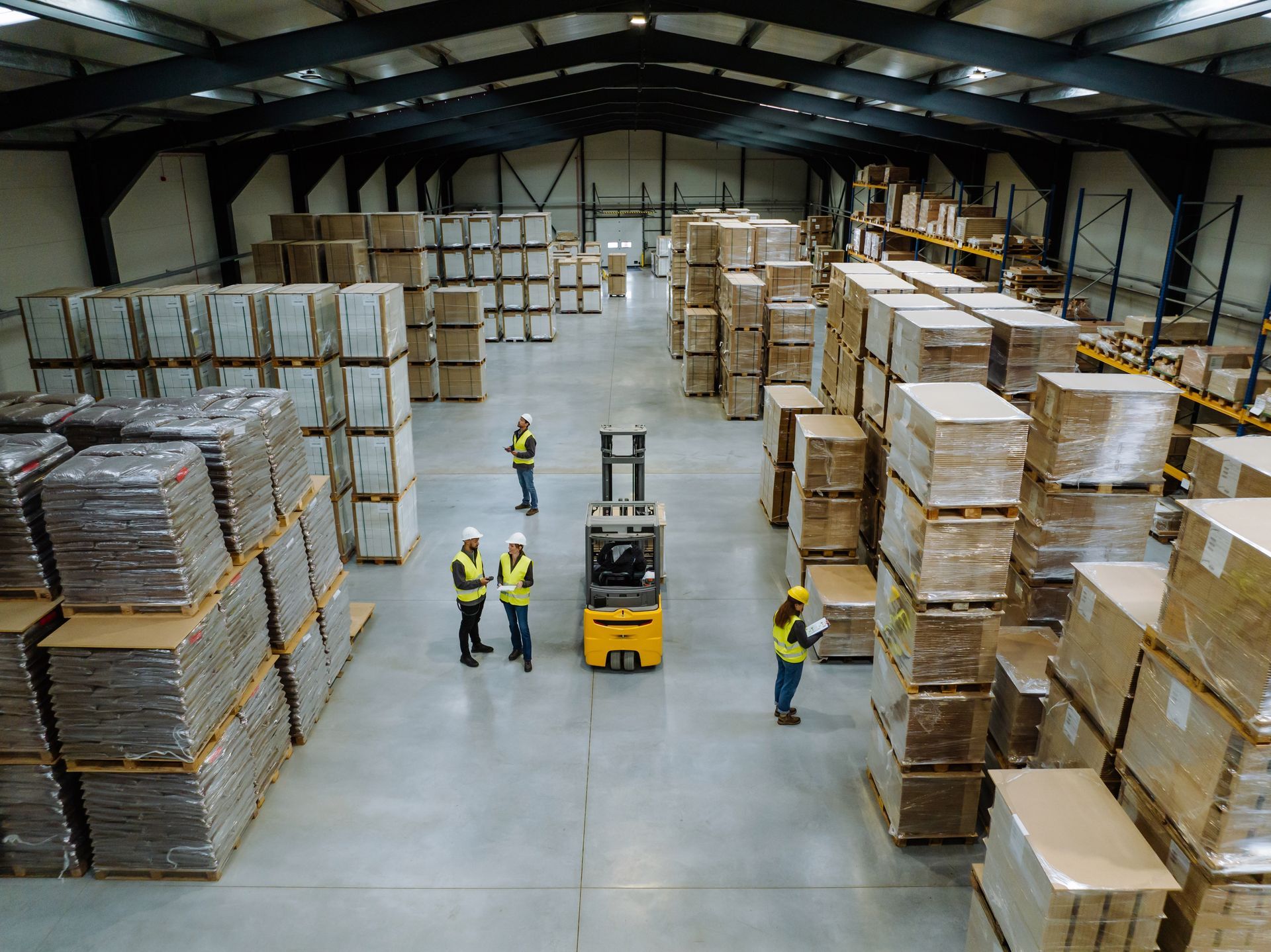 Warehouse workers in safety vests stand near a forklift in a large storage facility filled with stacks of palletized boxes.