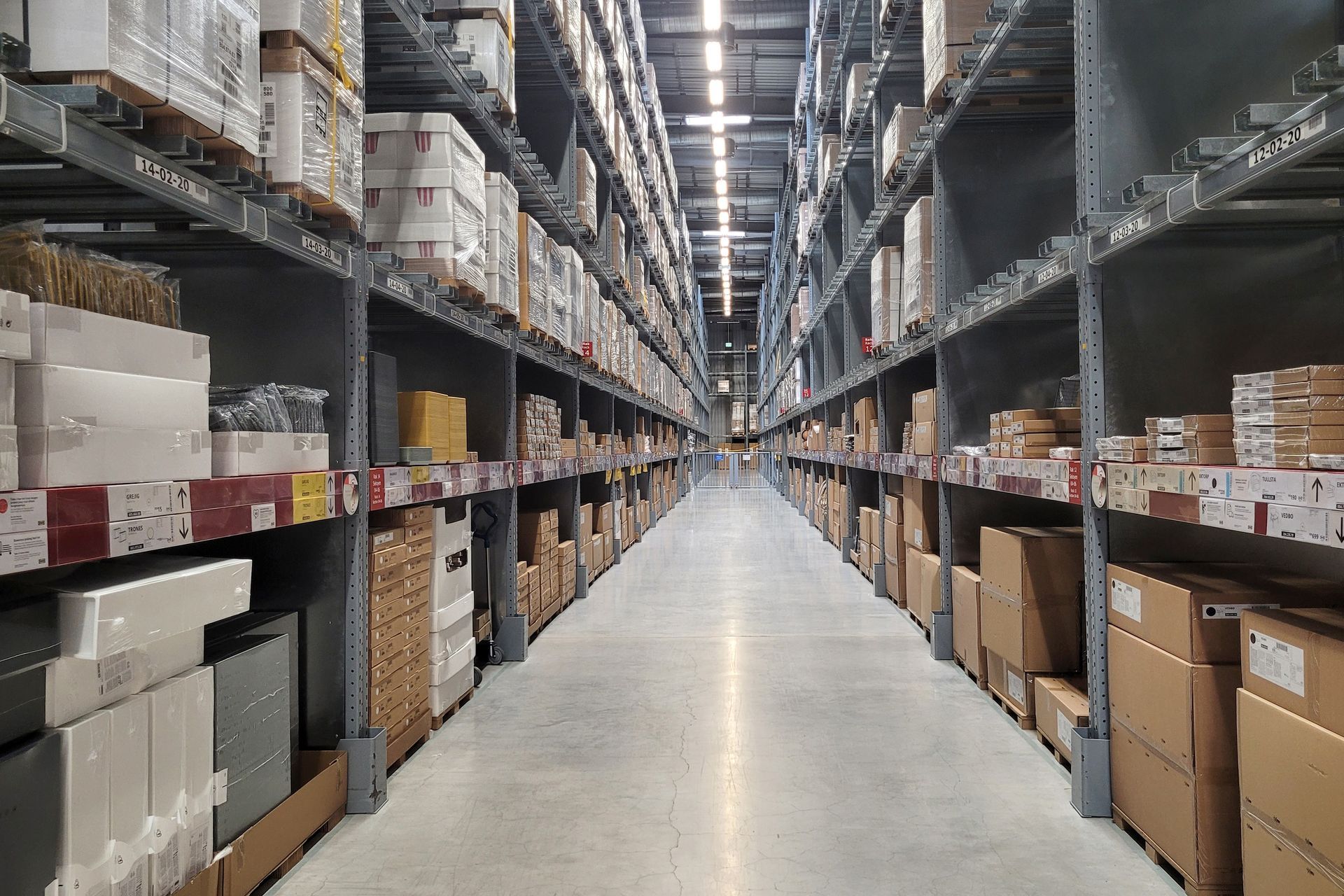 A high-ceiling warehouse aisle with metal shelving units filled with various cardboard boxes and retail inventory.