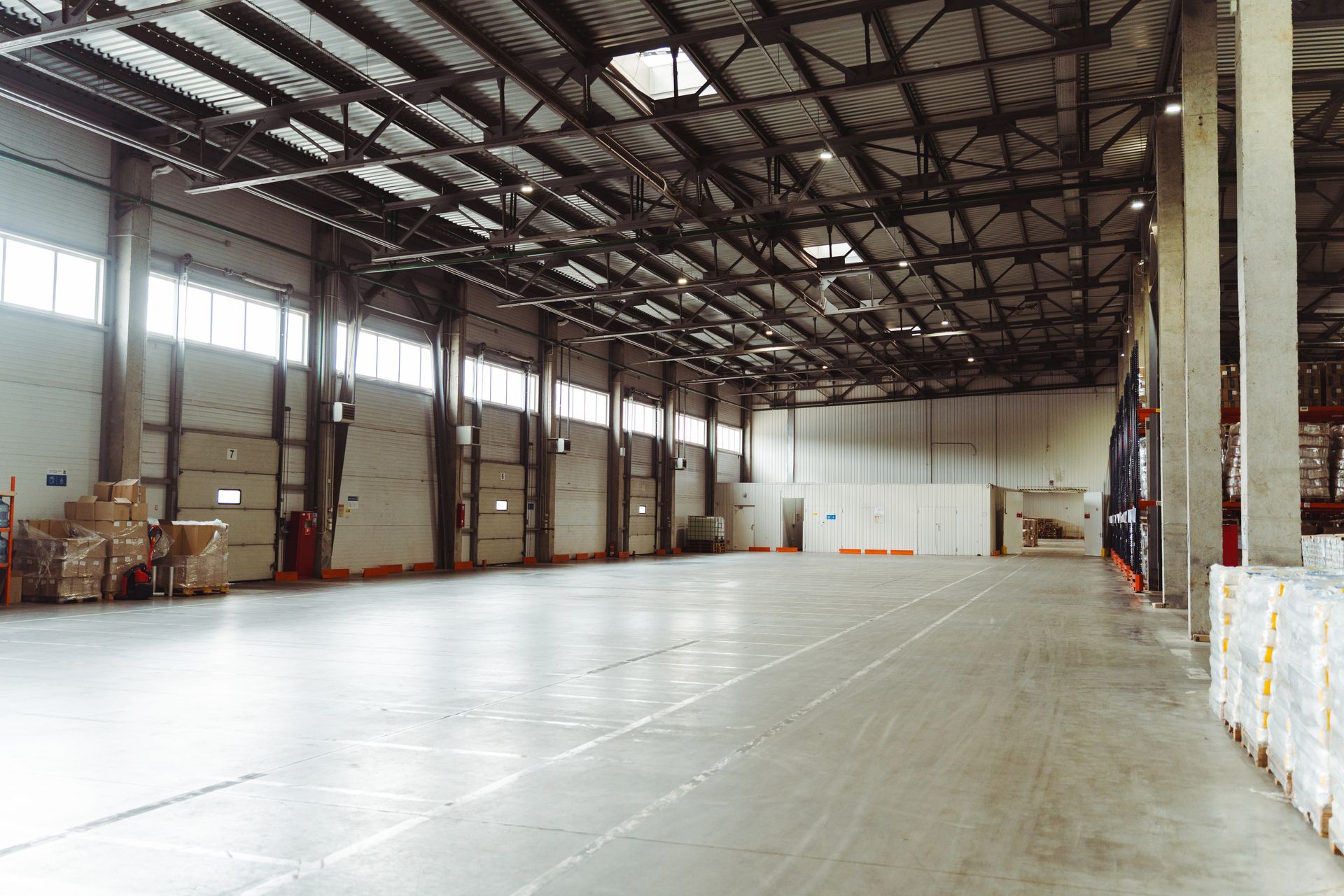 A wide, empty warehouse with concrete floors, high ceilings with metal trusses, and rows of windows along the wall.