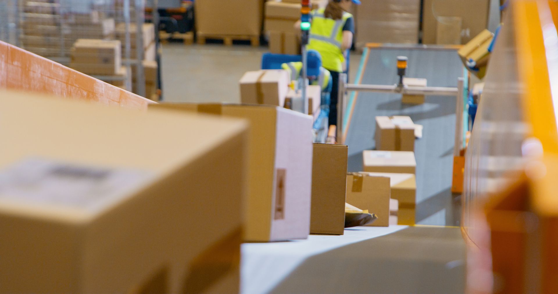 Cardboard boxes move along a conveyor belt in a warehouse, with a worker in a high-visibility vest visible in the back.