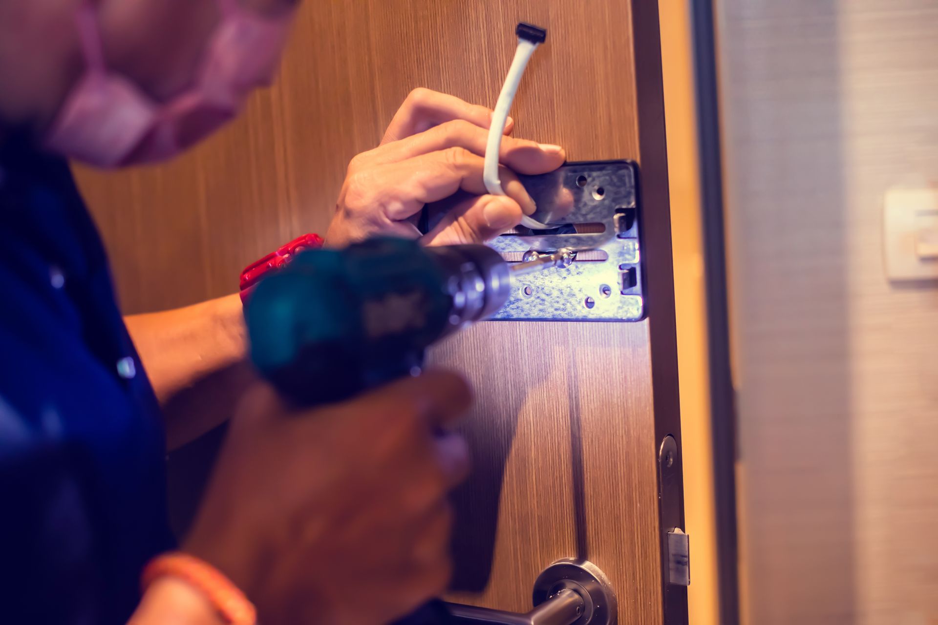 A person wearing a face mask uses a power drill to install a metal hardware plate onto a wooden door.