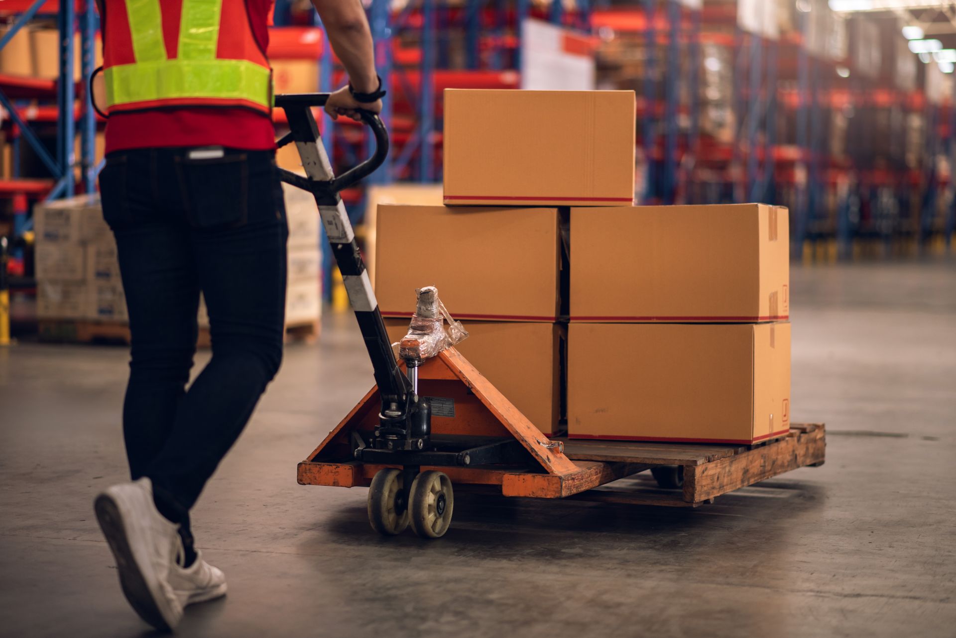 Warehouse worker in a high-visibility vest pulls a pallet jack loaded with cardboard boxes through a warehouse aisle.