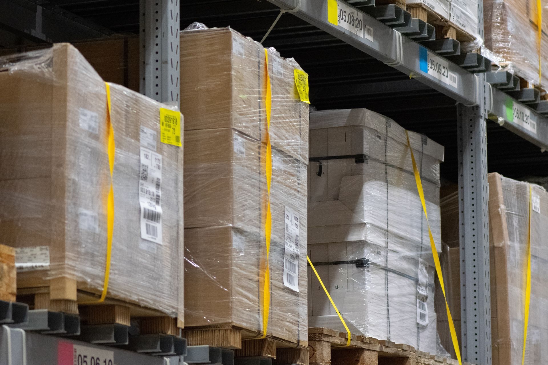 Cardboard boxes stacked on wooden pallets, wrapped in plastic, and stored on industrial warehouse metal shelving.
