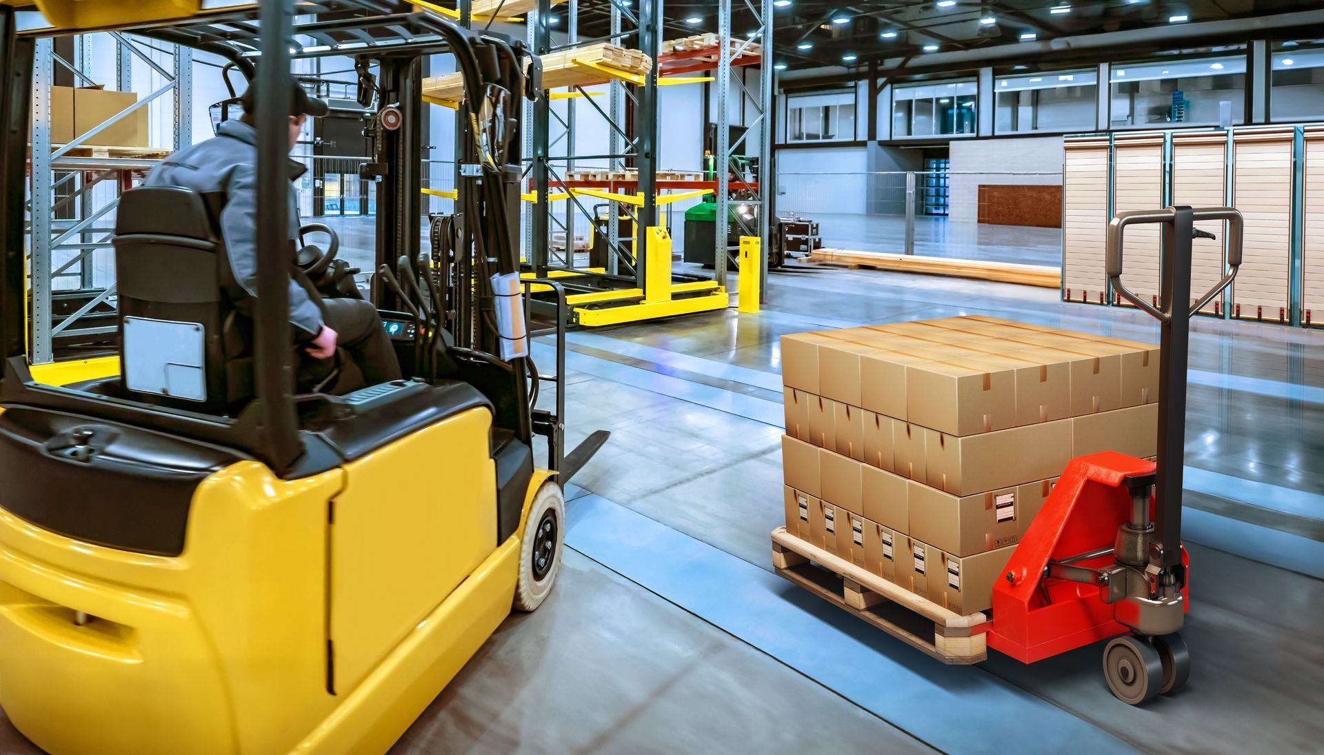 A person driving a yellow forklift near a red electric pallet jack loaded with cardboard boxes in a warehouse.