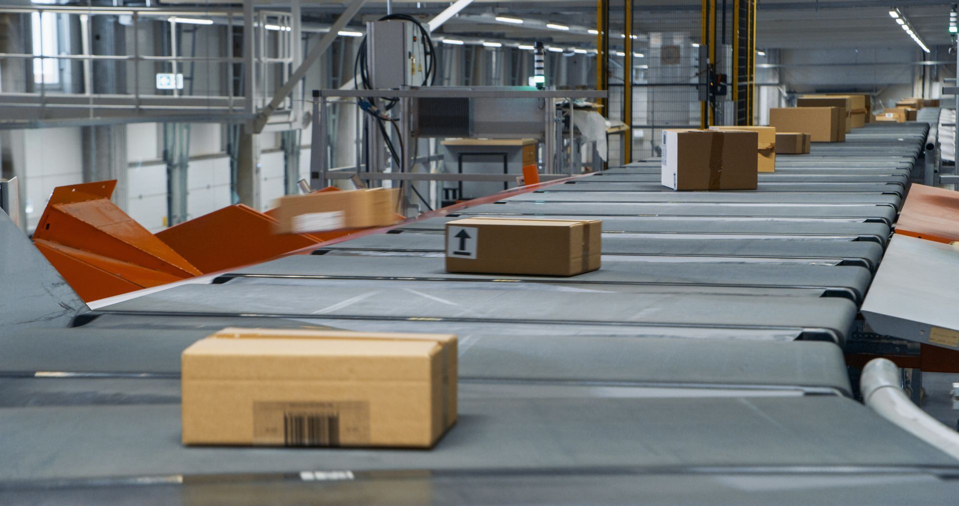 Cardboard shipping boxes move along a mechanized conveyor belt in a large, industrial warehouse facility.