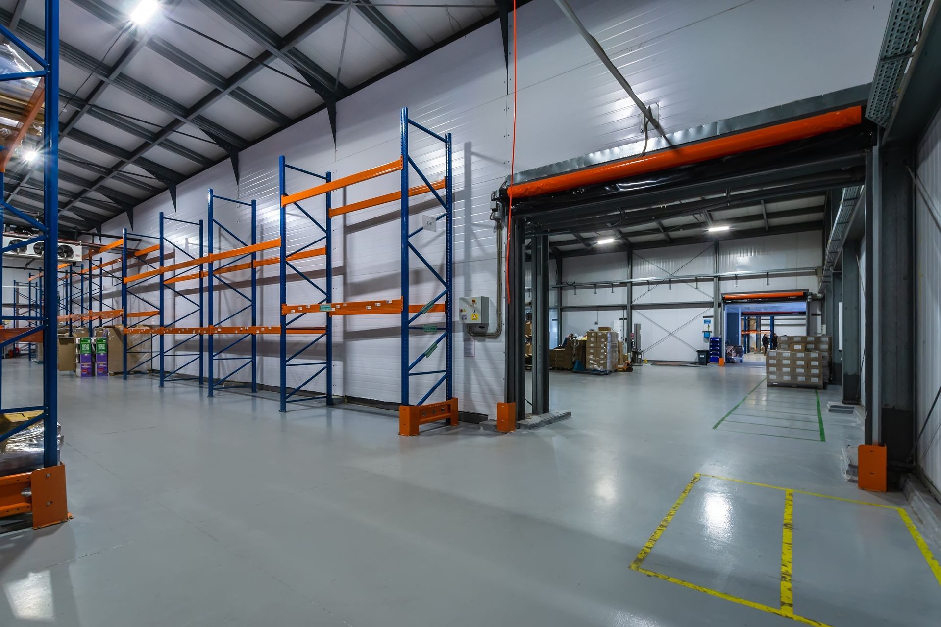 A spacious, well-lit warehouse with blue and orange metal storage racks along the walls and marked floor pathways.