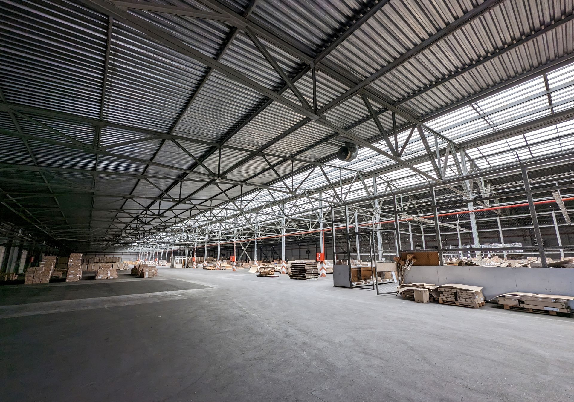 Spacious, open-plan industrial warehouse with a high metal-truss ceiling, gray concrete floor, and stacks of materials.