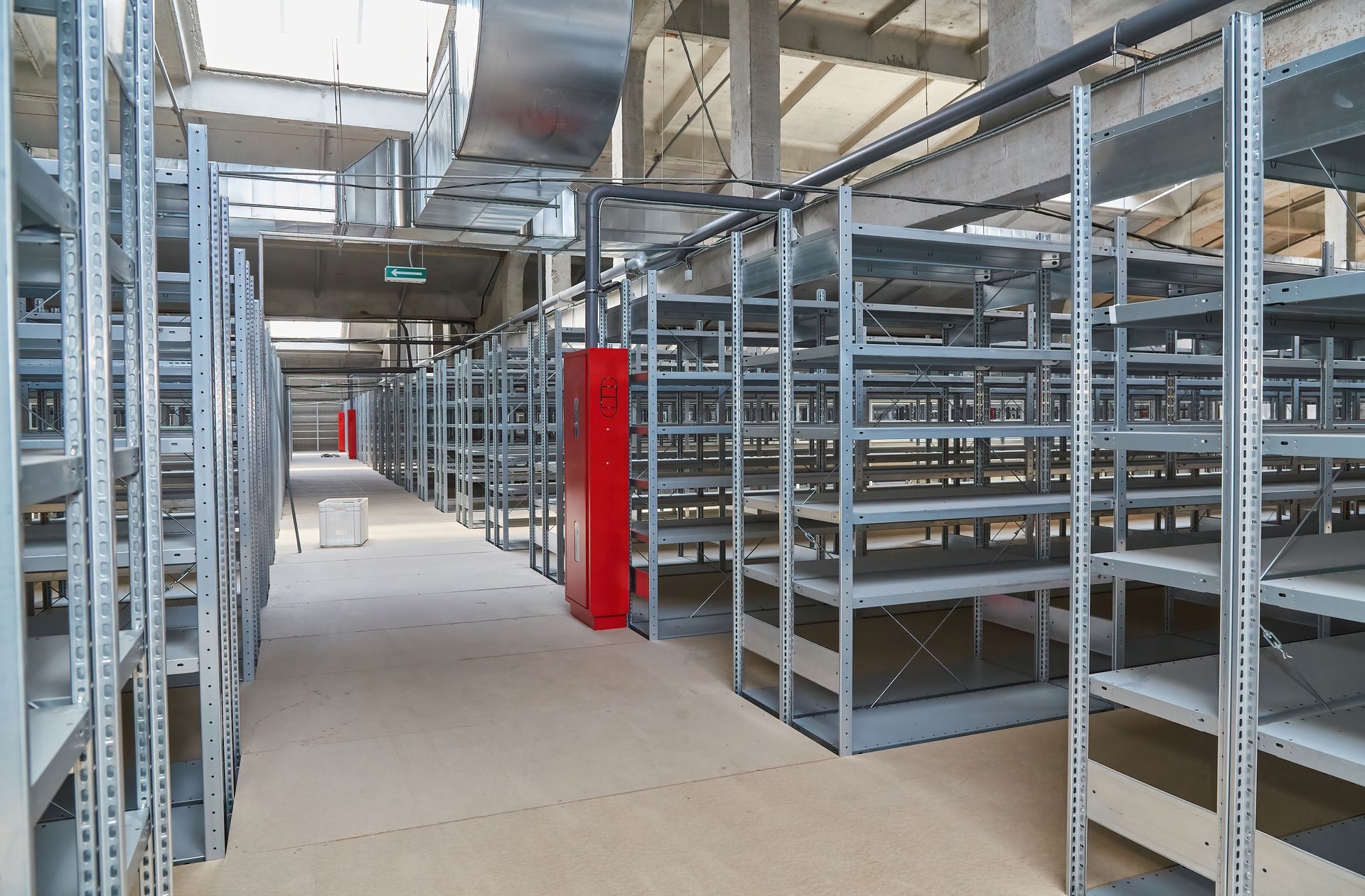 Rows of empty metal shelving units inside a large warehouse with visible ventilation ducts and concrete pillars.