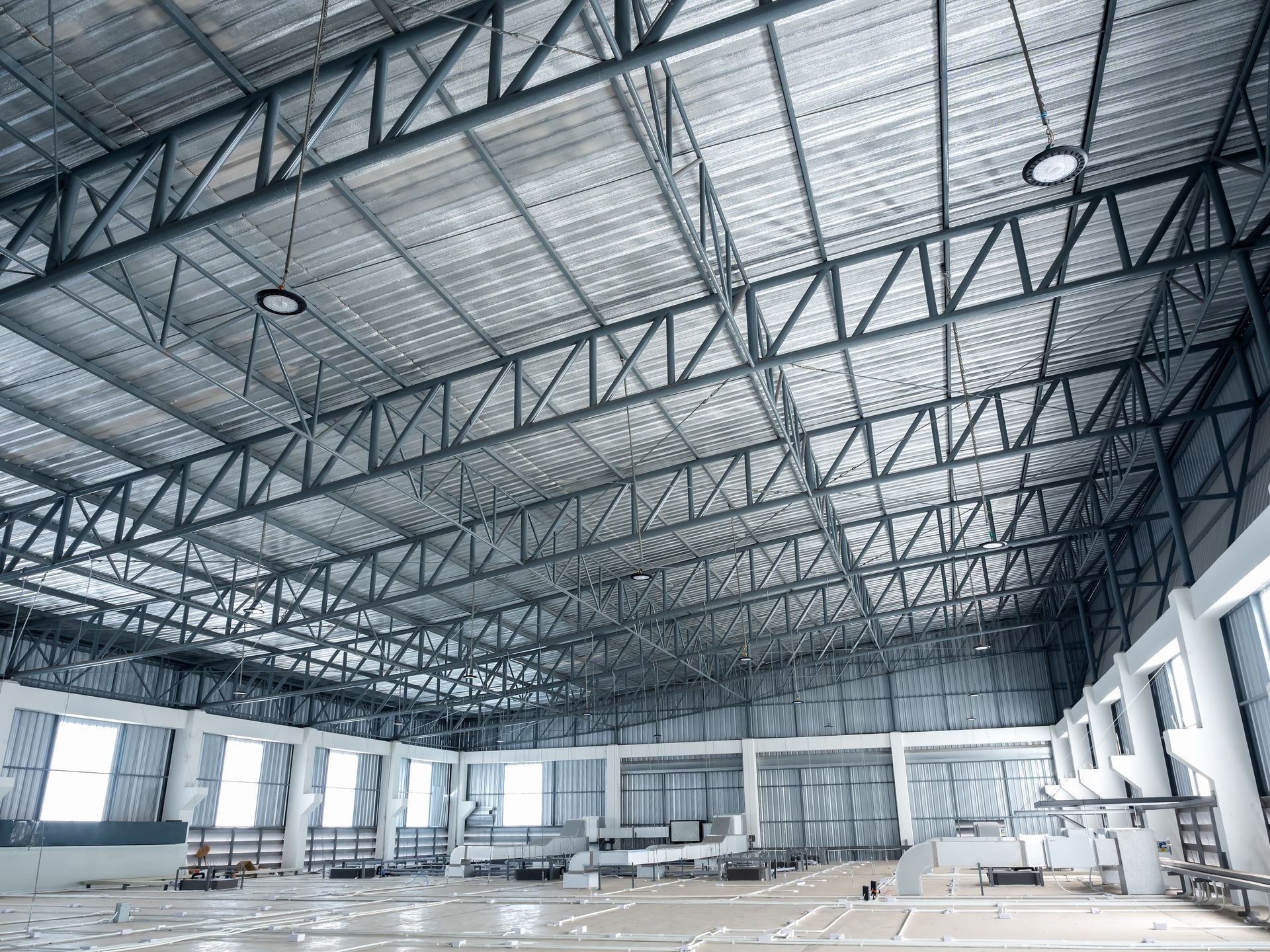 Interior view of a large, unfinished industrial warehouse with exposed metal truss ceilings, concrete floors, and walls.