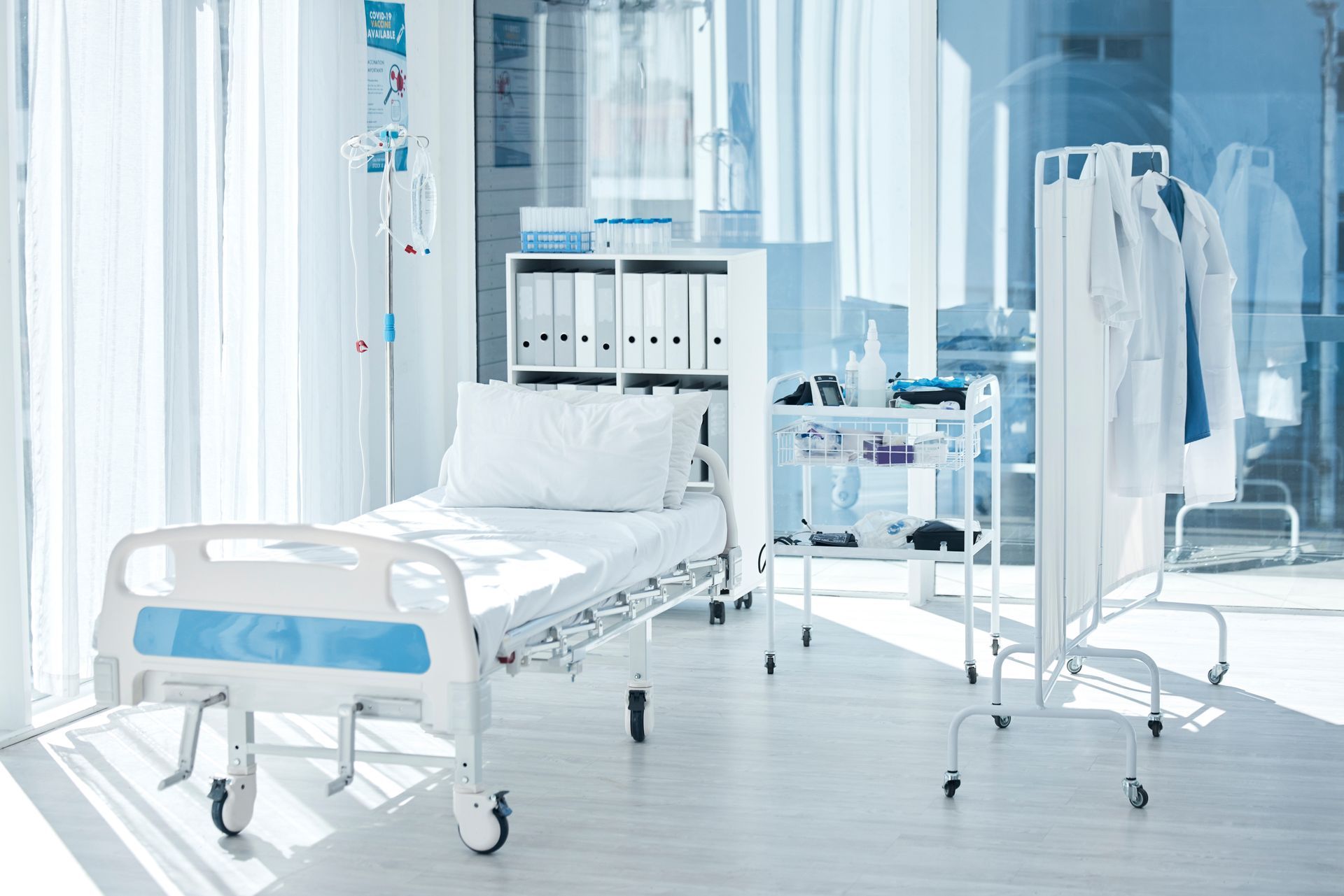 A clean, bright hospital room featuring a bed, a shelf with binders, medical equipment, and a mobile rack with lab coats.