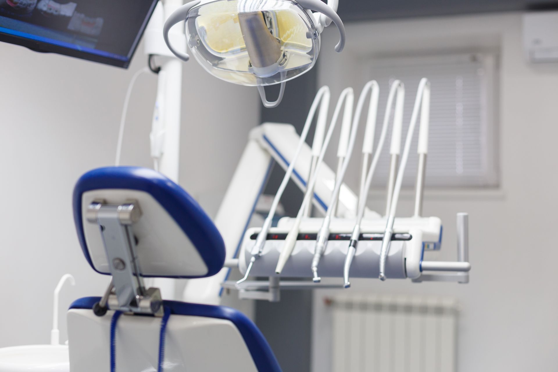 A dental chair with blue upholstery facing a station of hanging handheld dental instruments and an overhead lamp.