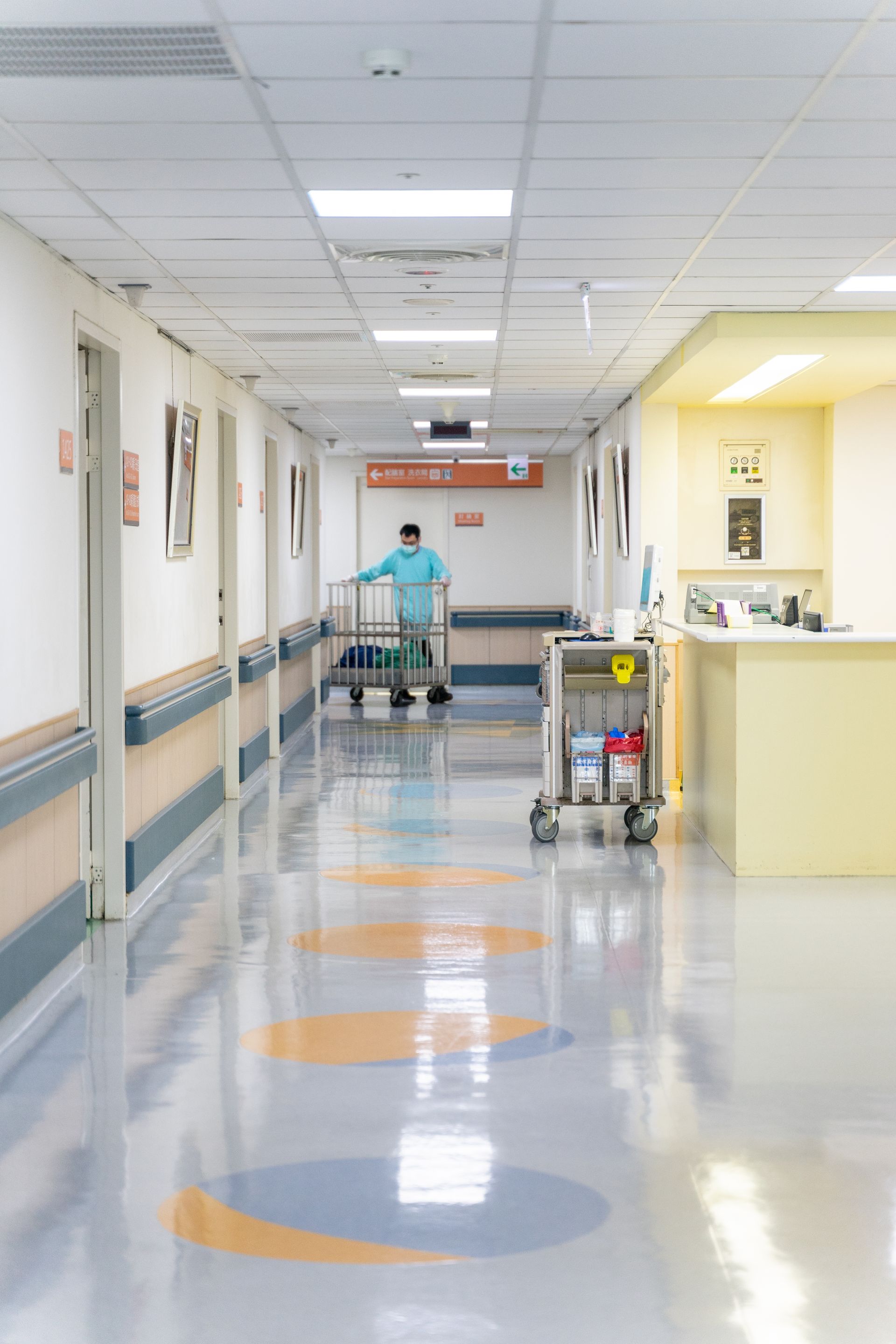 A hallway in a hospital with a staff member pushing a cart, and a medical cart sitting by a yellow nursing station.