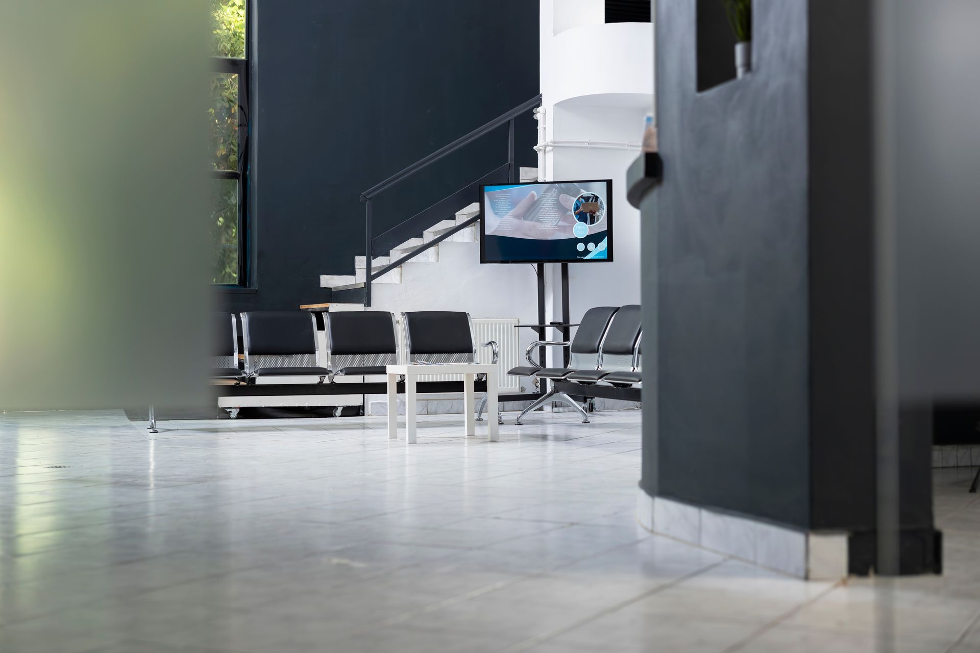 Modern waiting room with black seating, a central TV monitor on a stand, and a light-colored tiled floor.