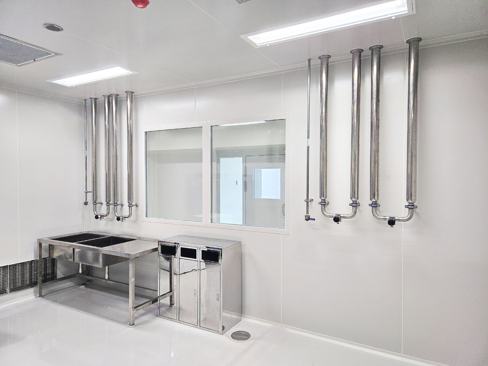 Clean room interior with sterile white walls, stainless steel laboratory tables, and vertical metal piping.