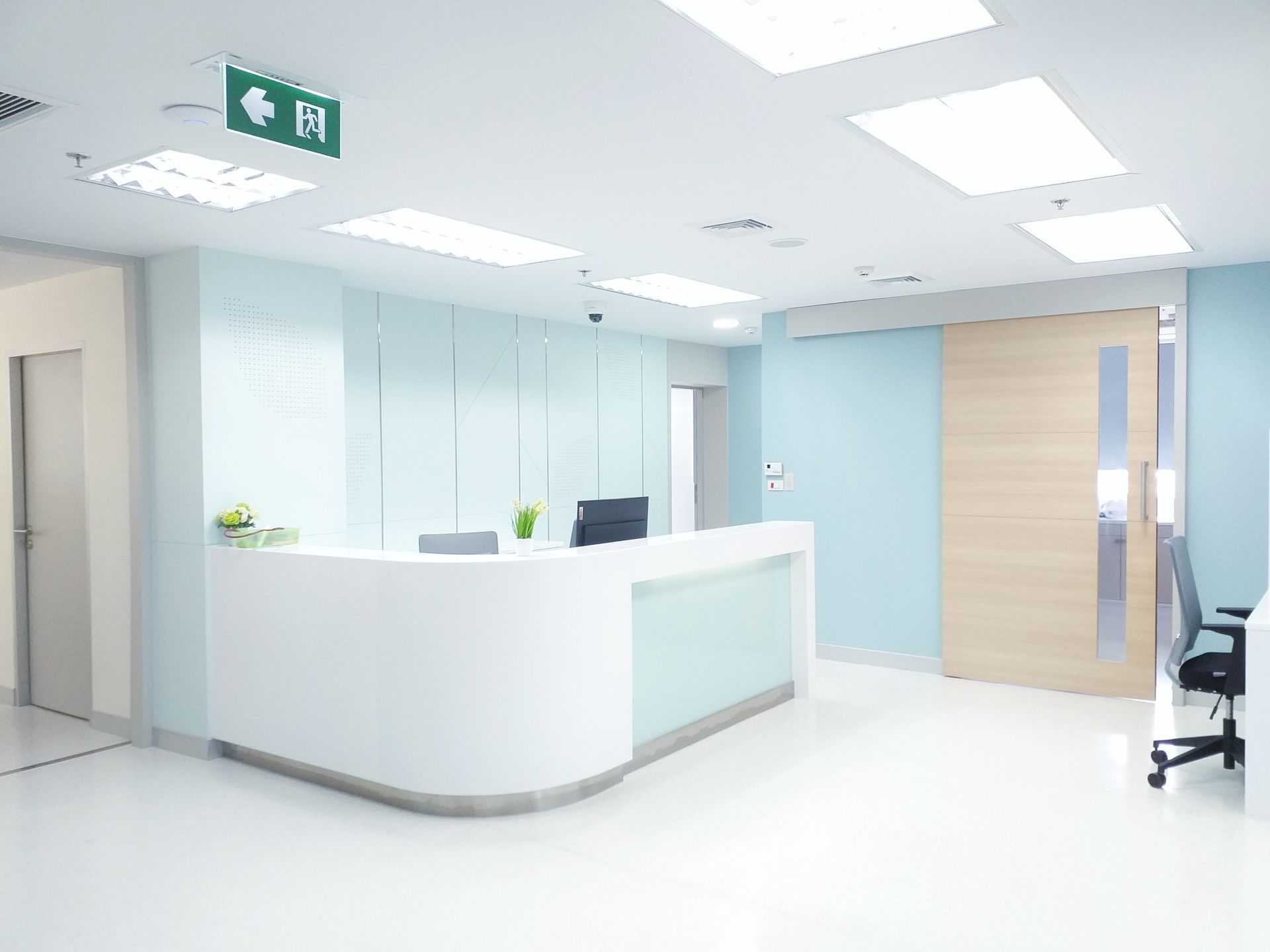 A modern medical reception area featuring a white curved desk, light blue walls, a wooden door, and overhead lighting.