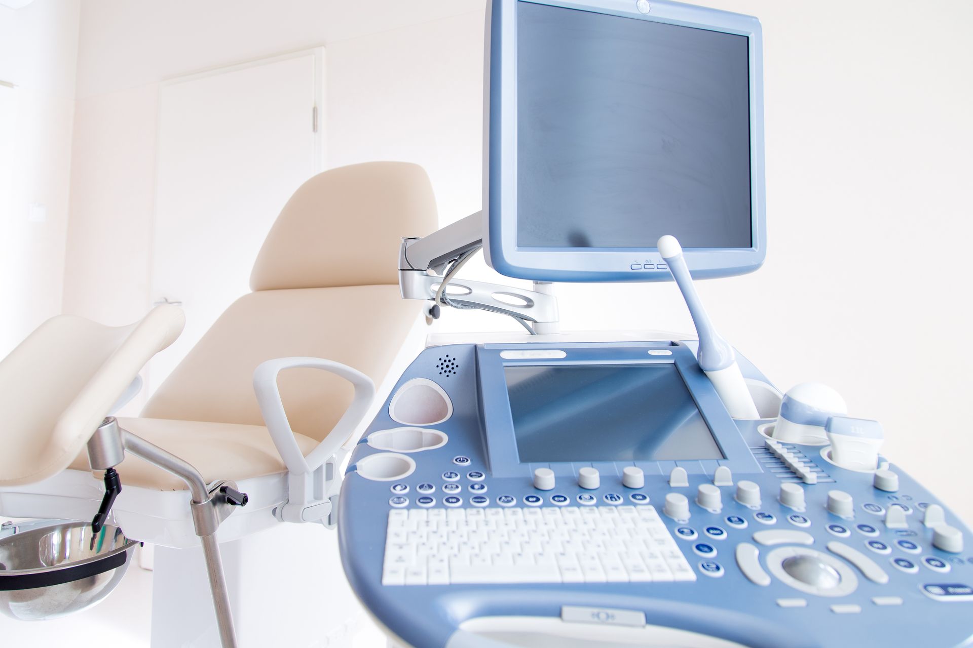 An ultrasound machine sits in a bright, clean medical examination room next to an adjustable medical chair.
