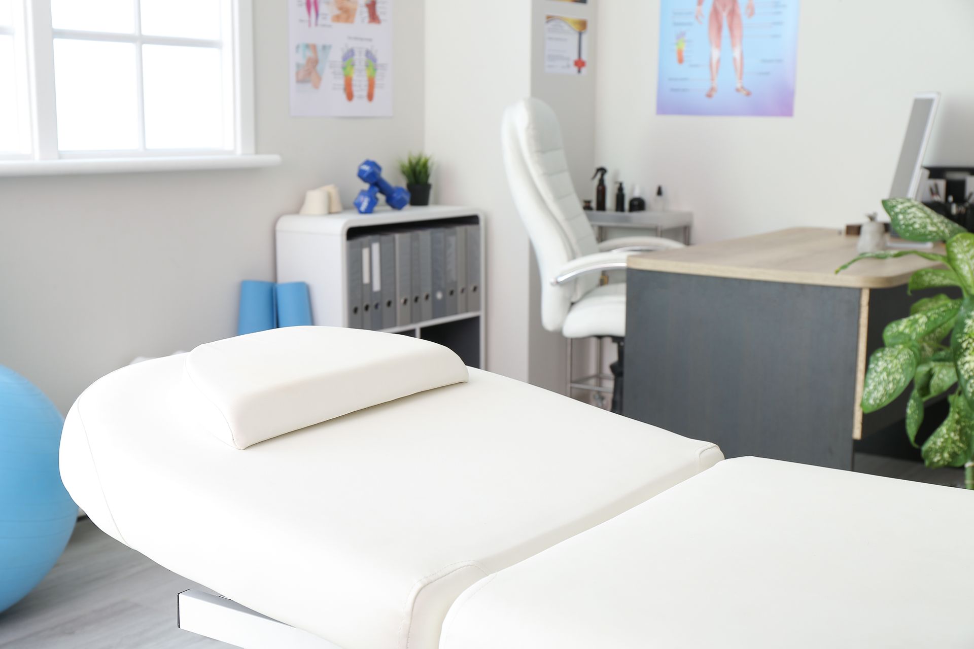 A white massage table in a bright physical therapy office with a desk, office chair, and anatomical charts on the wall.