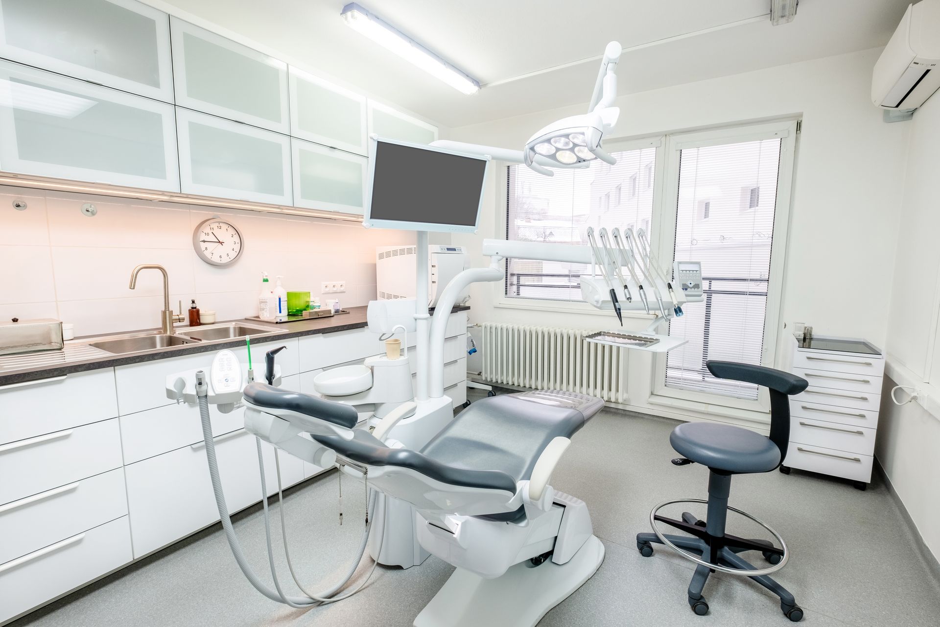 A bright, modern dental examination room with a gray patient chair, an adjustable stool, and medical equipment.